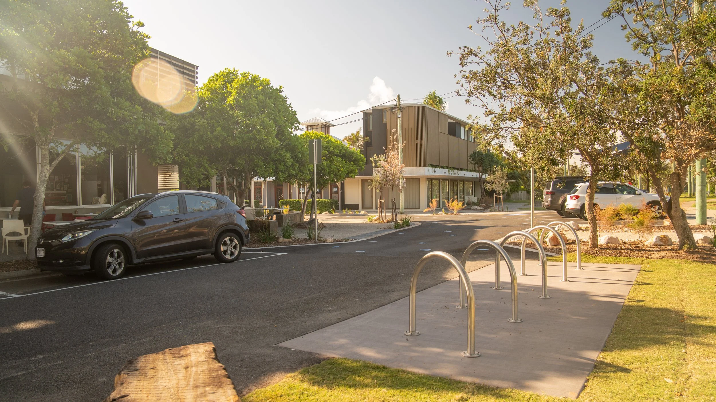 A parking lot with a black car parked near a sidewalk, several trees, and modern buildings in the background on a sunny day.