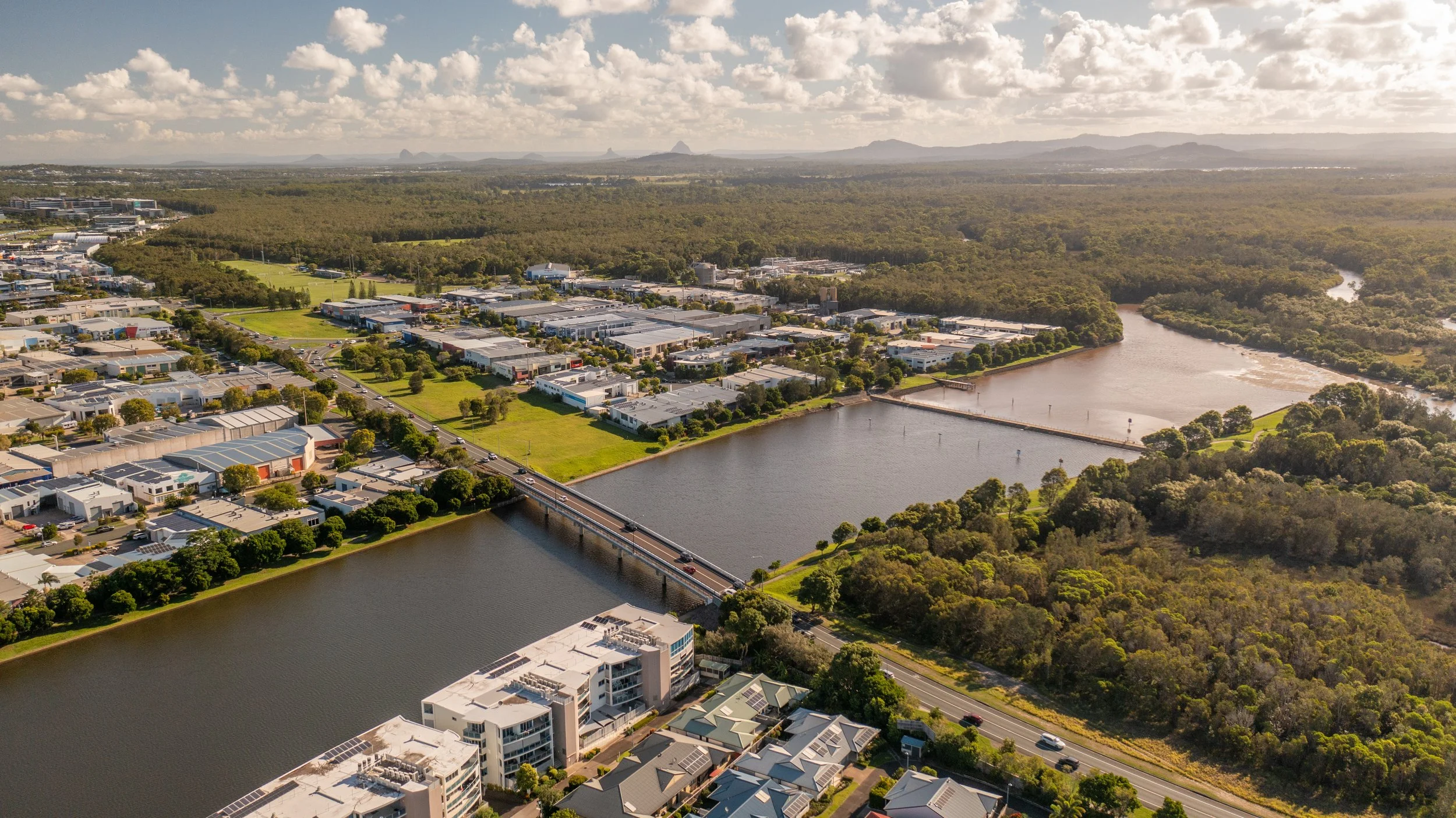 An aerial view of a city with a river running through it, bridges crossing the water, surrounded by residential and commercial buildings, green parks, and wooded areas under a partly cloudy sky.