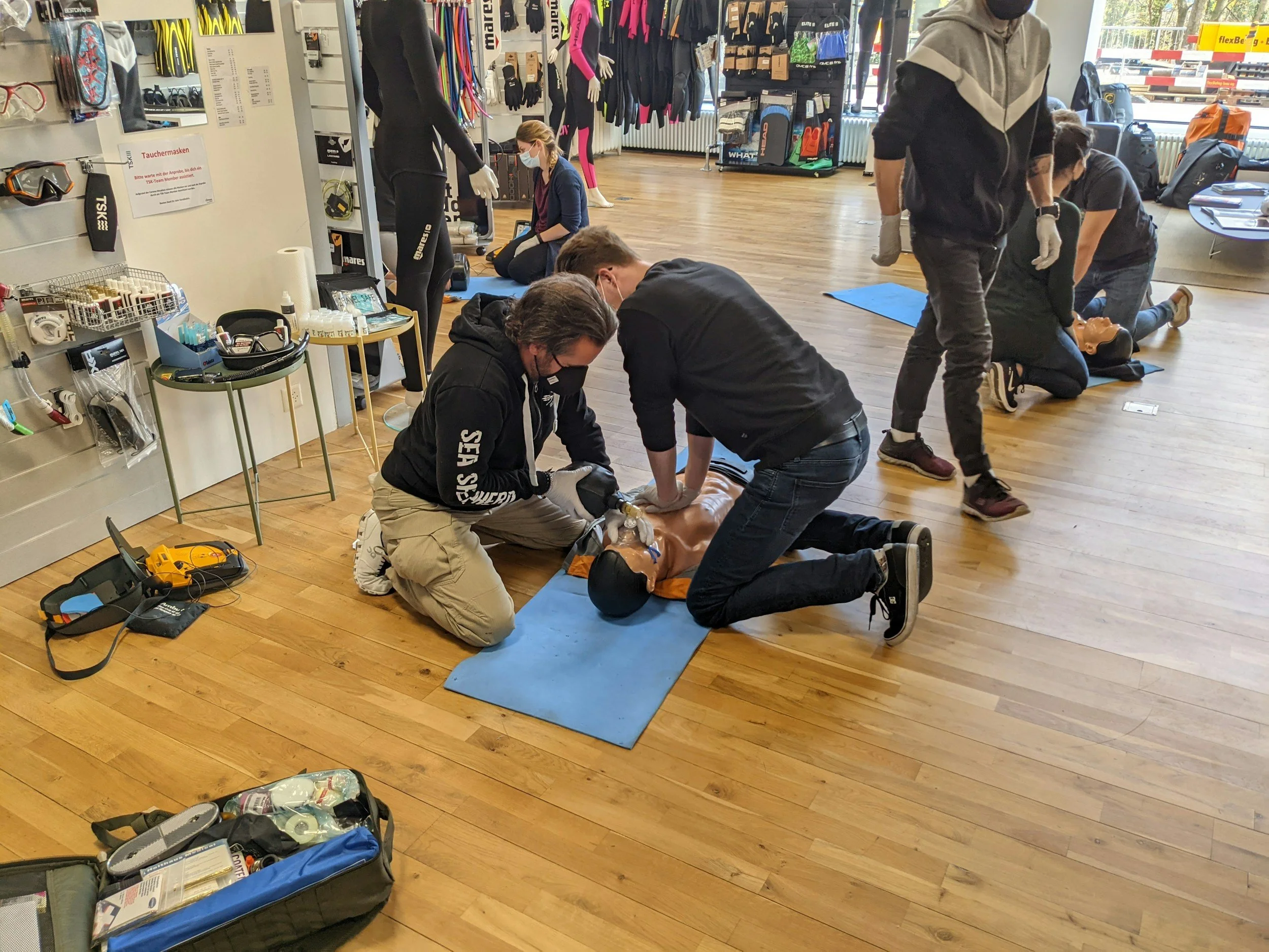 People practicing CPR on mannequins indoors on wooden floor with medical kits and diving gear visible.