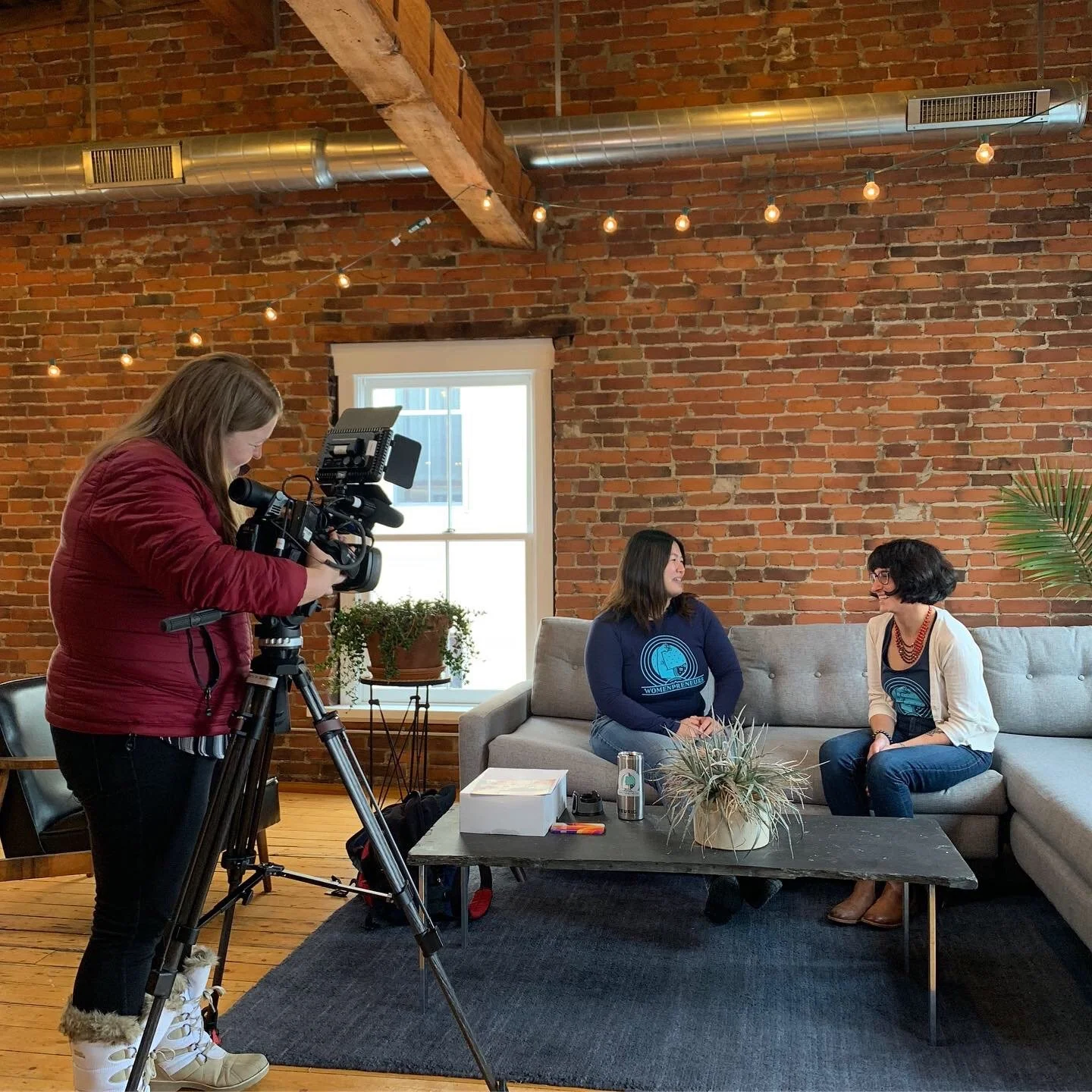 Two women seated on a gray couch in a room with exposed brick walls, being recorded by a camera operator with a professional camera. There is a black wooden table in front with a potted plant, a box, and some items on it. A window and some potted plants are visible in the background.