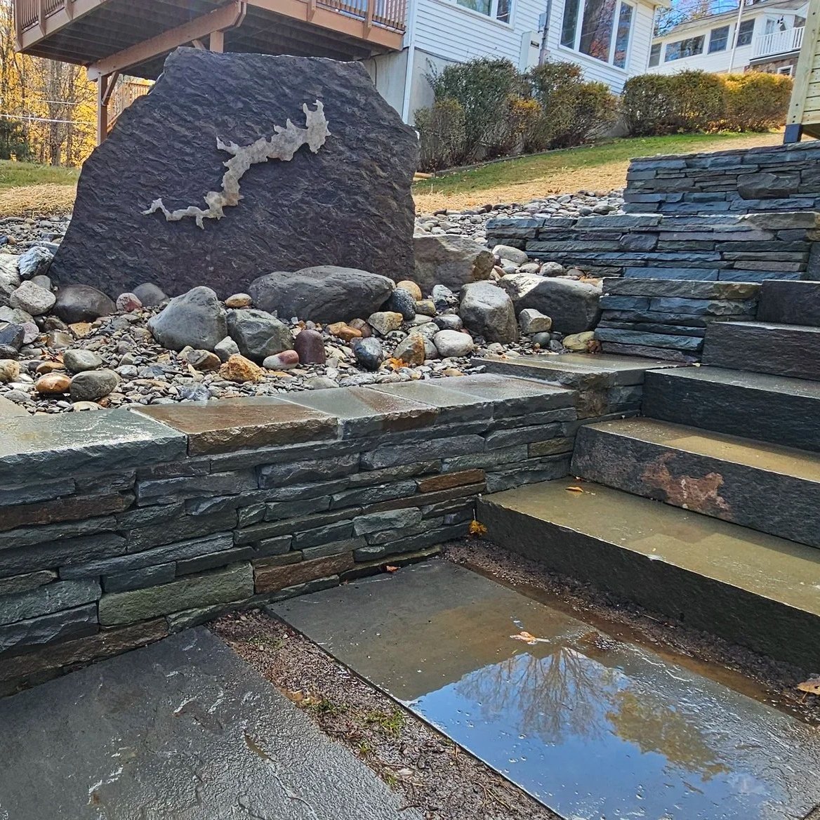 A landscaped backyard with stone steps, a stone retaining wall, a large stone with a map of Michigan, and a dry creek bed with rocks.