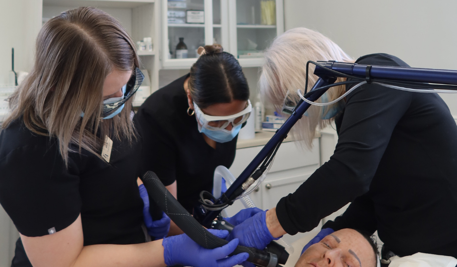 Three trained clinicians wearing protective eyewear and gloves perform a CO₂ laser resurfacing treatment on a patient in a clinical environment.