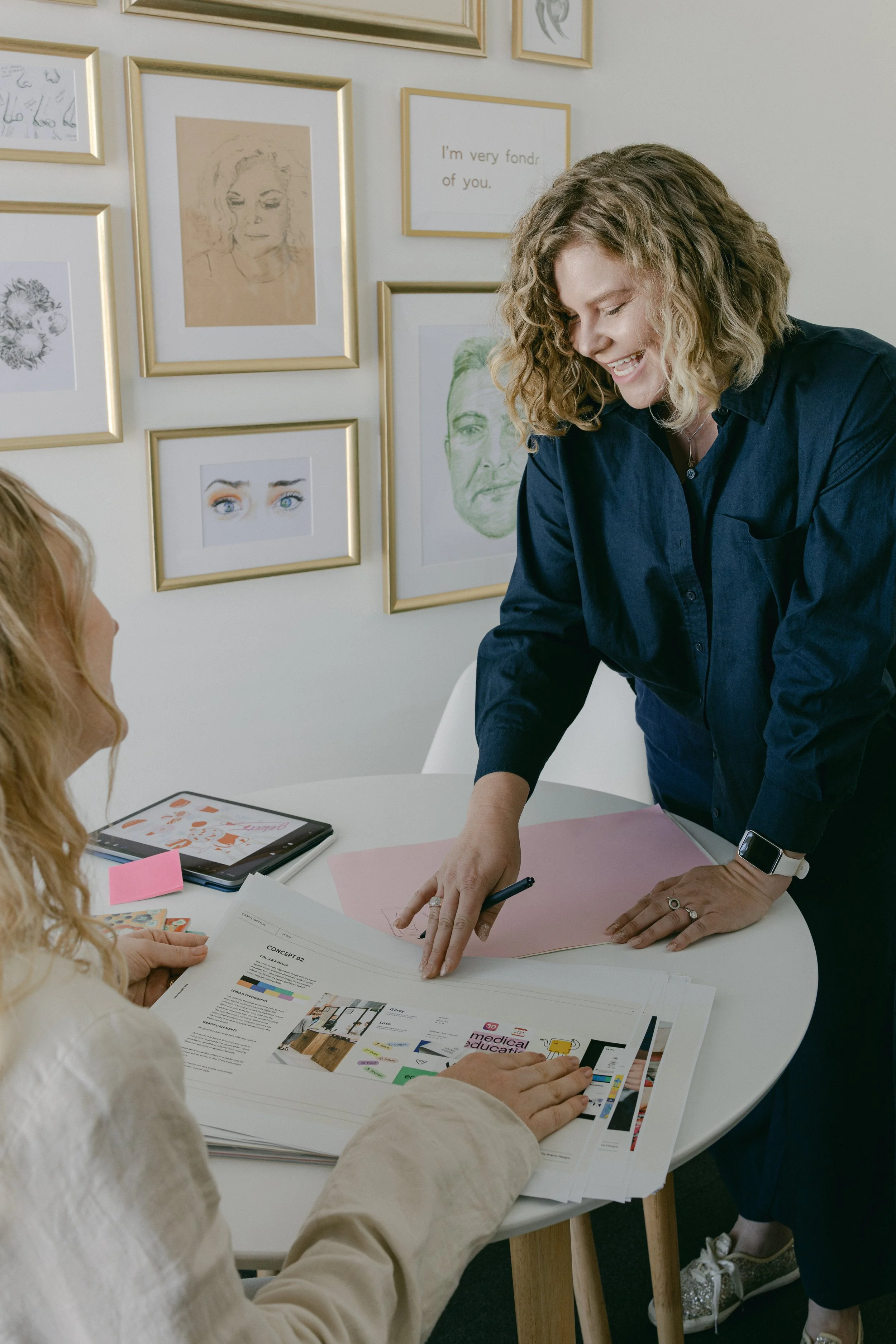 Two women collaborating at a round table in a modern office. One woman is standing, pointing at printed materials, while the other is seated, looking at the documents. The wall behind them displays various framed art and quotes.