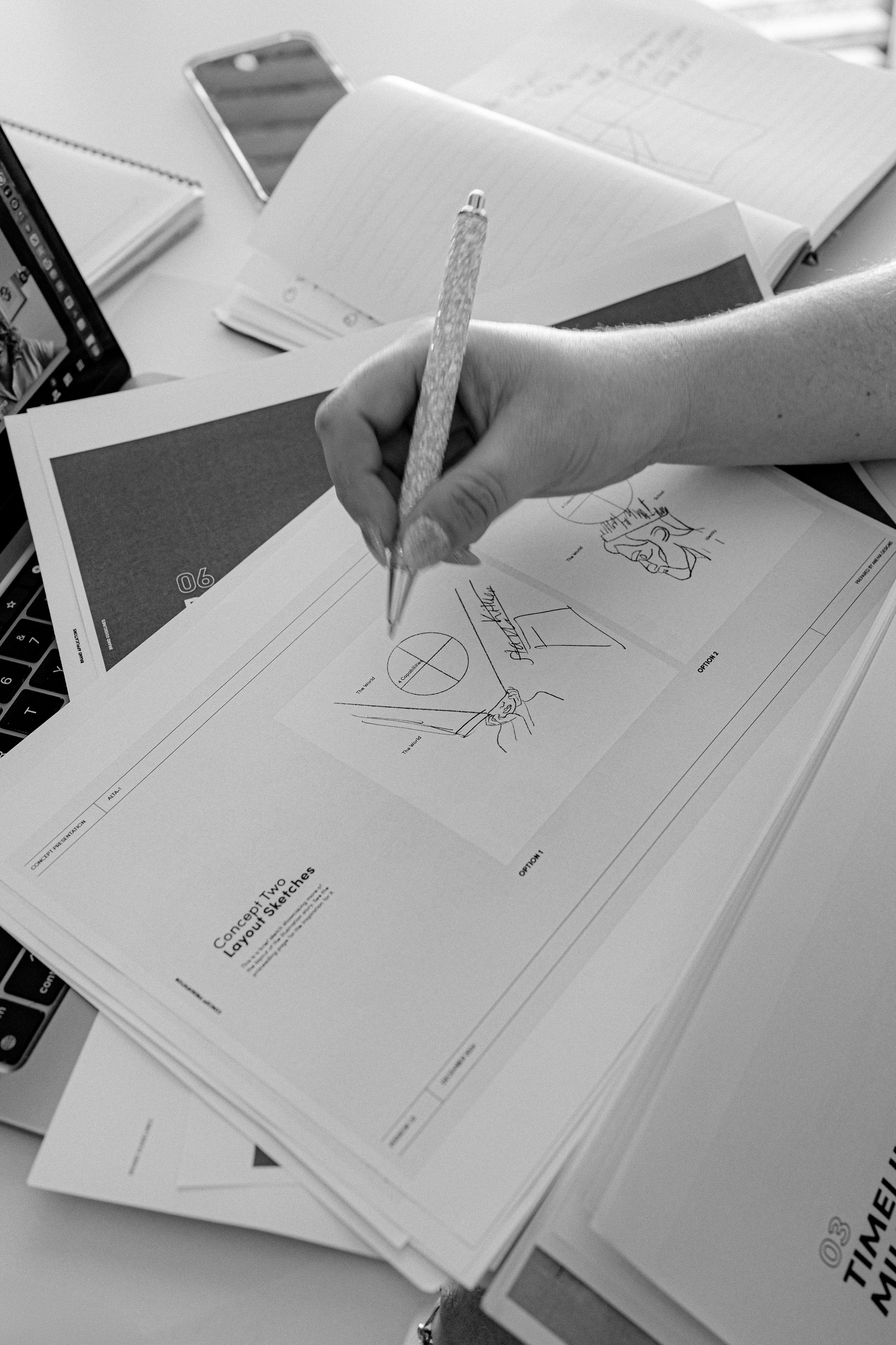 A person is sketching on concept layout sketches at a desk, with notebooks, a smartphone, and a laptop nearby.