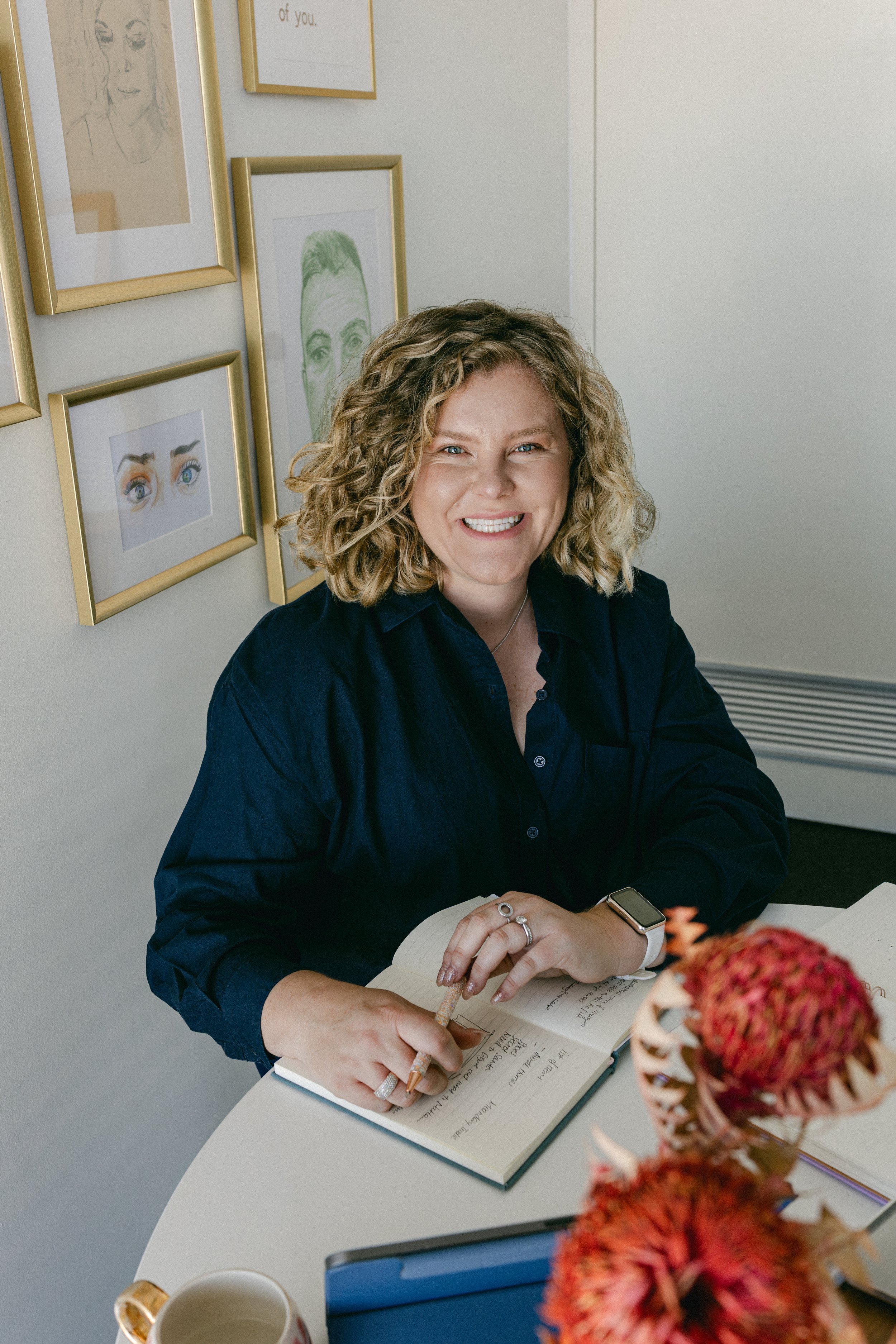 A smiling woman with curly blonde hair, wearing a dark blue shirt, sitting at a white table with an open notebook, pen, and a smartwatch on her wrist. She is in a room with white walls decorated with framed artwork, including illustrations of eyes and faces.