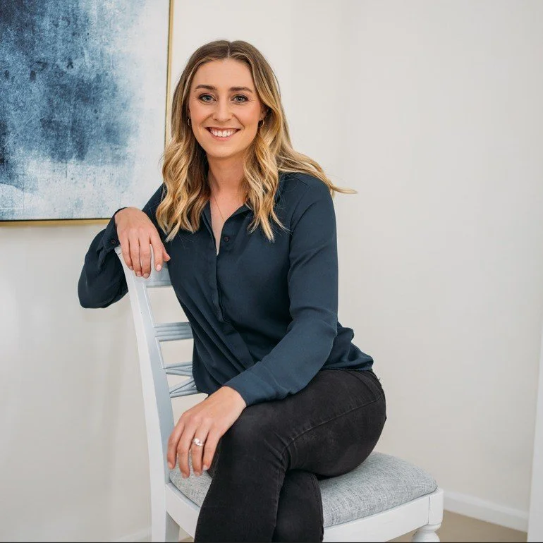 A woman with long blonde hair, wearing a navy blue blouse and black pants, sitting on a white chair with a light gray cushion, smiling indoors against a white wall with a blue artwork on it.