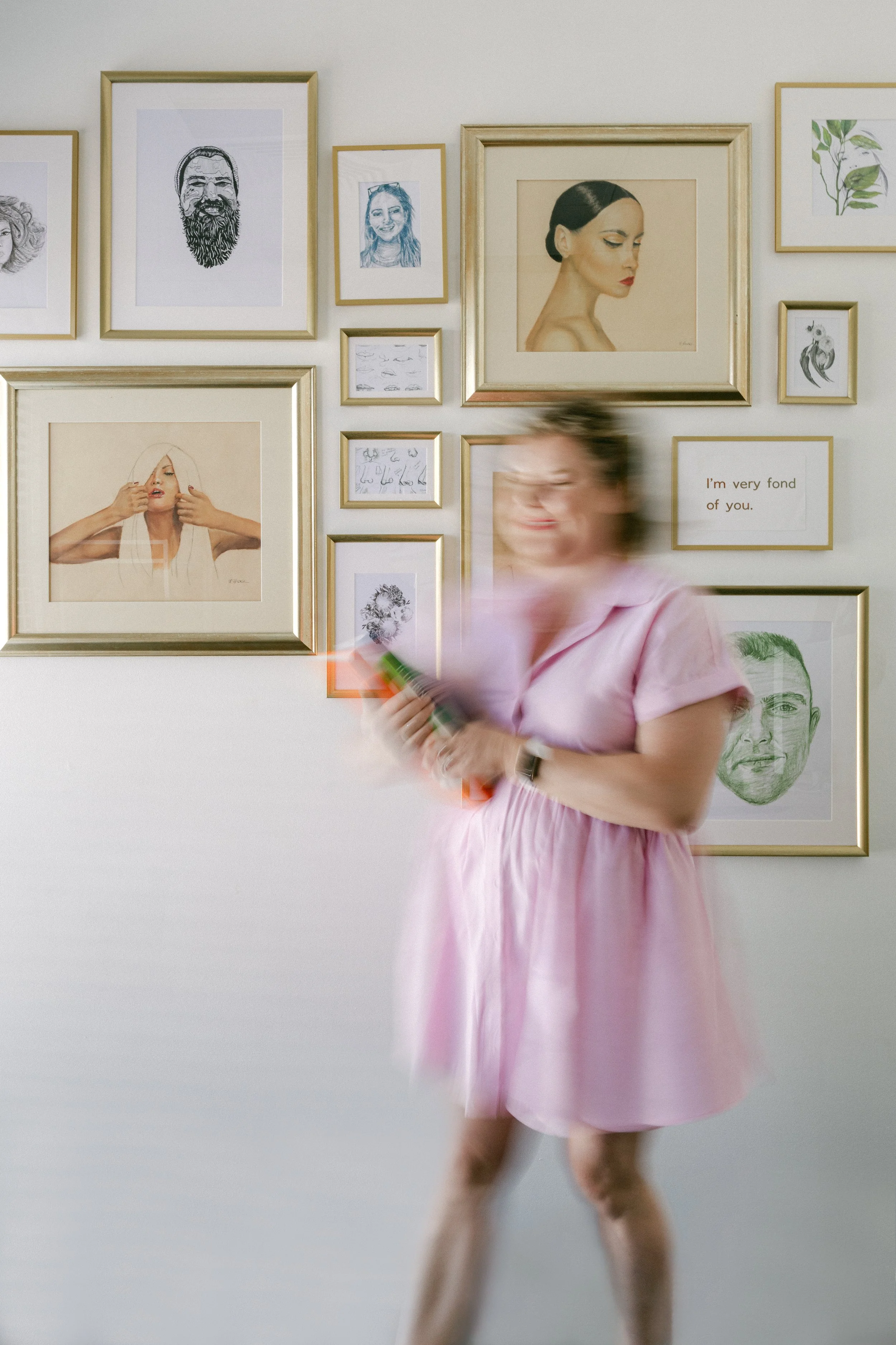 A woman in a pink dress is walking past a wall decorated with various framed pictures and artwork, including portraits, drawings, and a quote.