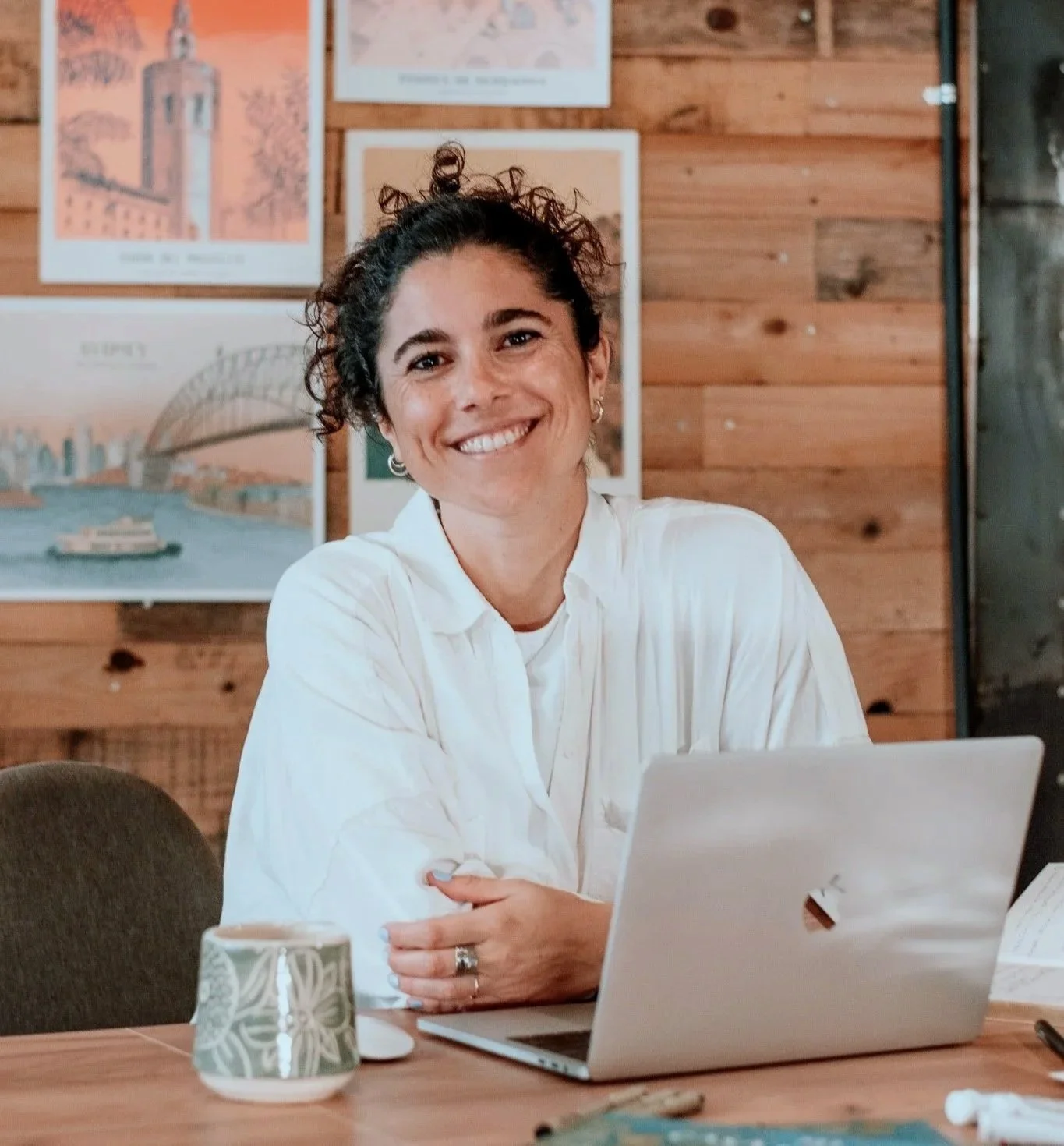 A woman with curly dark hair smiling, sitting at a wooden table with a silver laptop, mug, and papers, against a wooden wall with framed artwork.