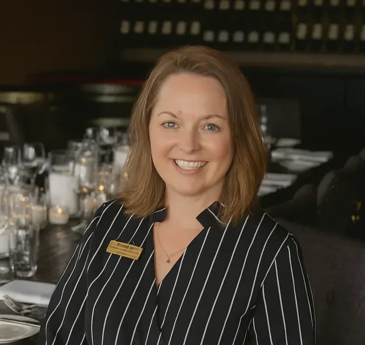 A smiling woman with shoulder-length light brown hair, wearing a black and white pinstripe uniform with a gold name tag, sitting at a table set with glassware and candles in a restaurant.