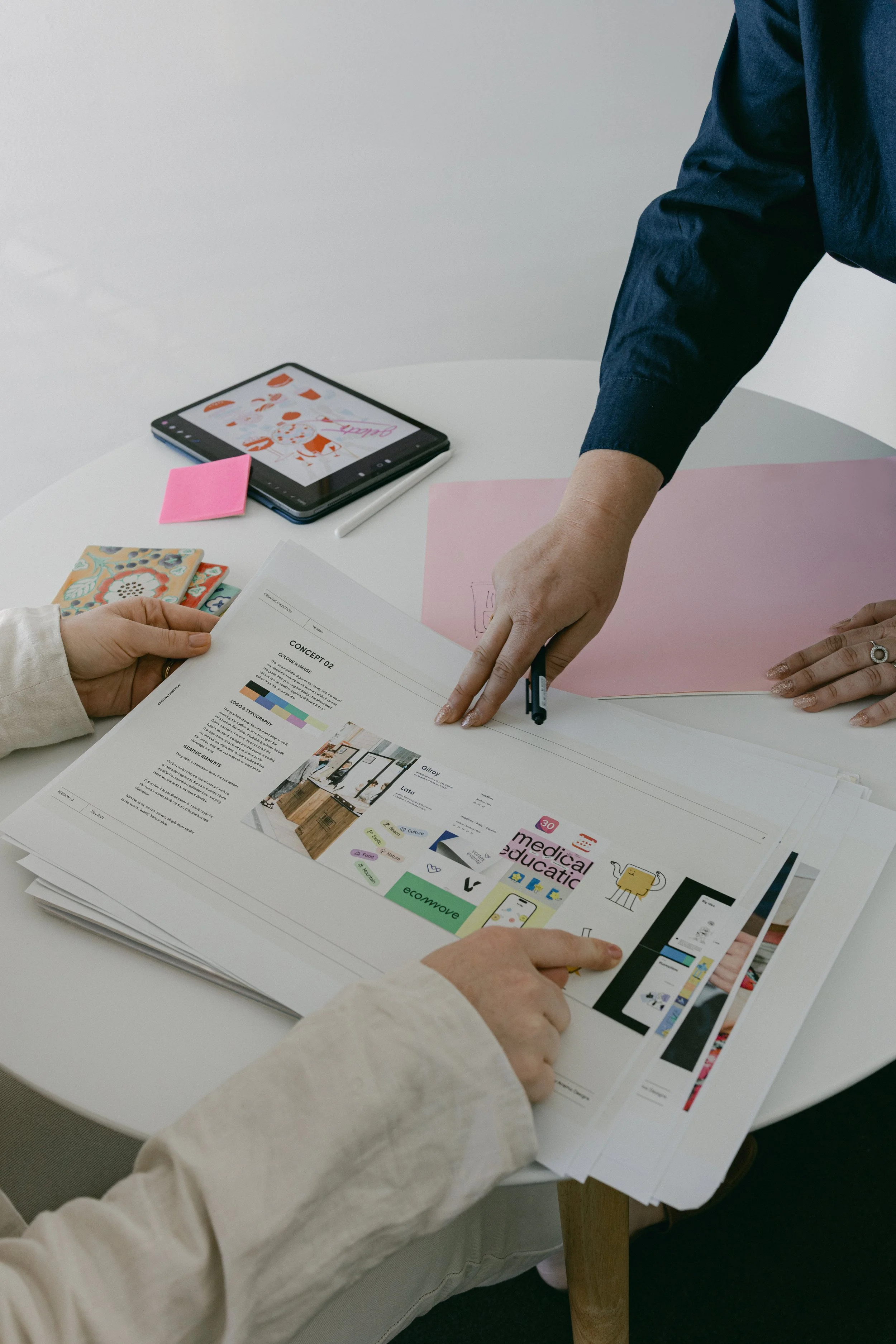 Two people working on a project at a white table with printed documents, a tablet, sticky notes, and colorful papers. One person points at a sheet of paper, while the other holds it.