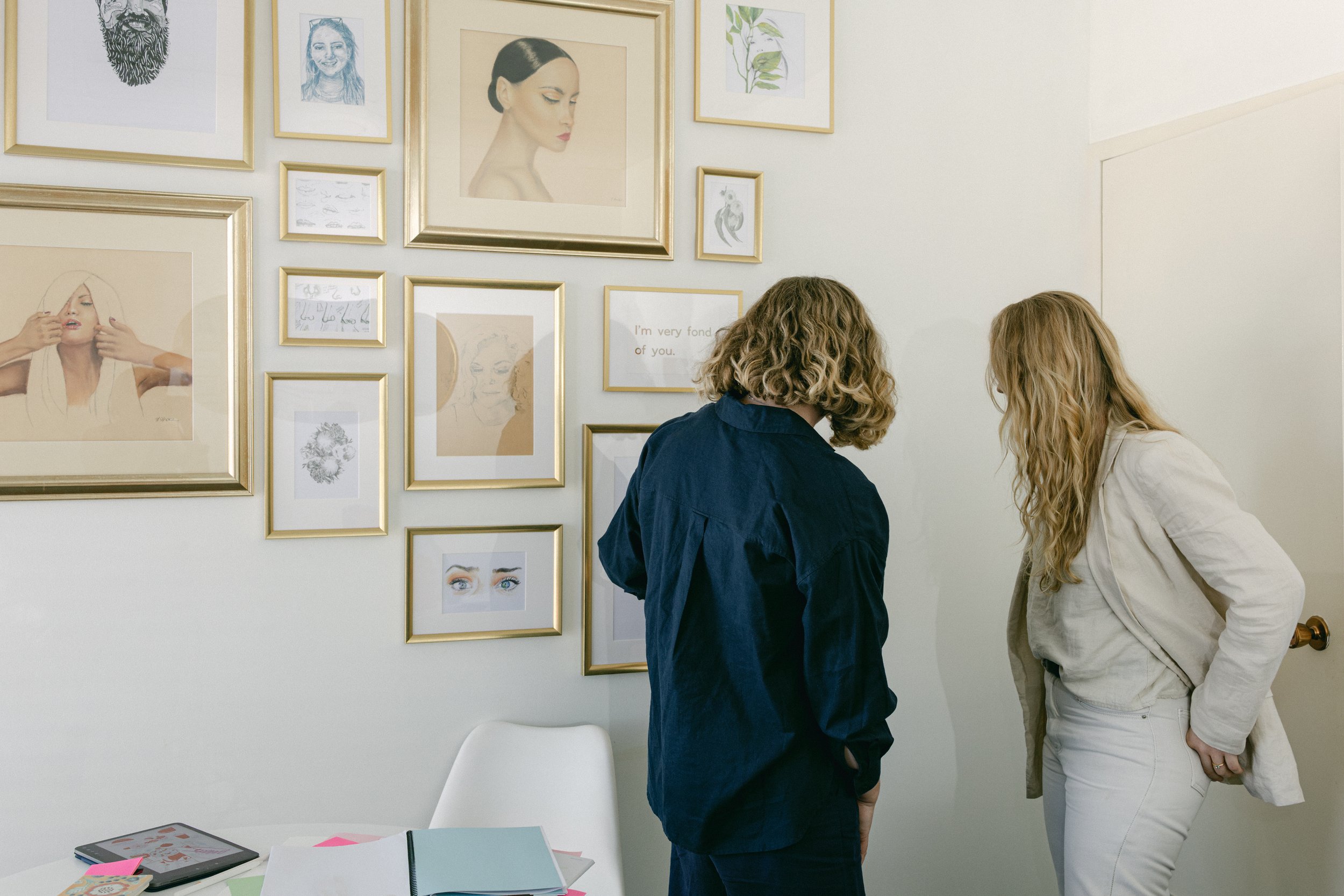 Two women standing and talking in a room with a white wall decorated with various framed pictures and drawings, and a table with notebooks and papers.