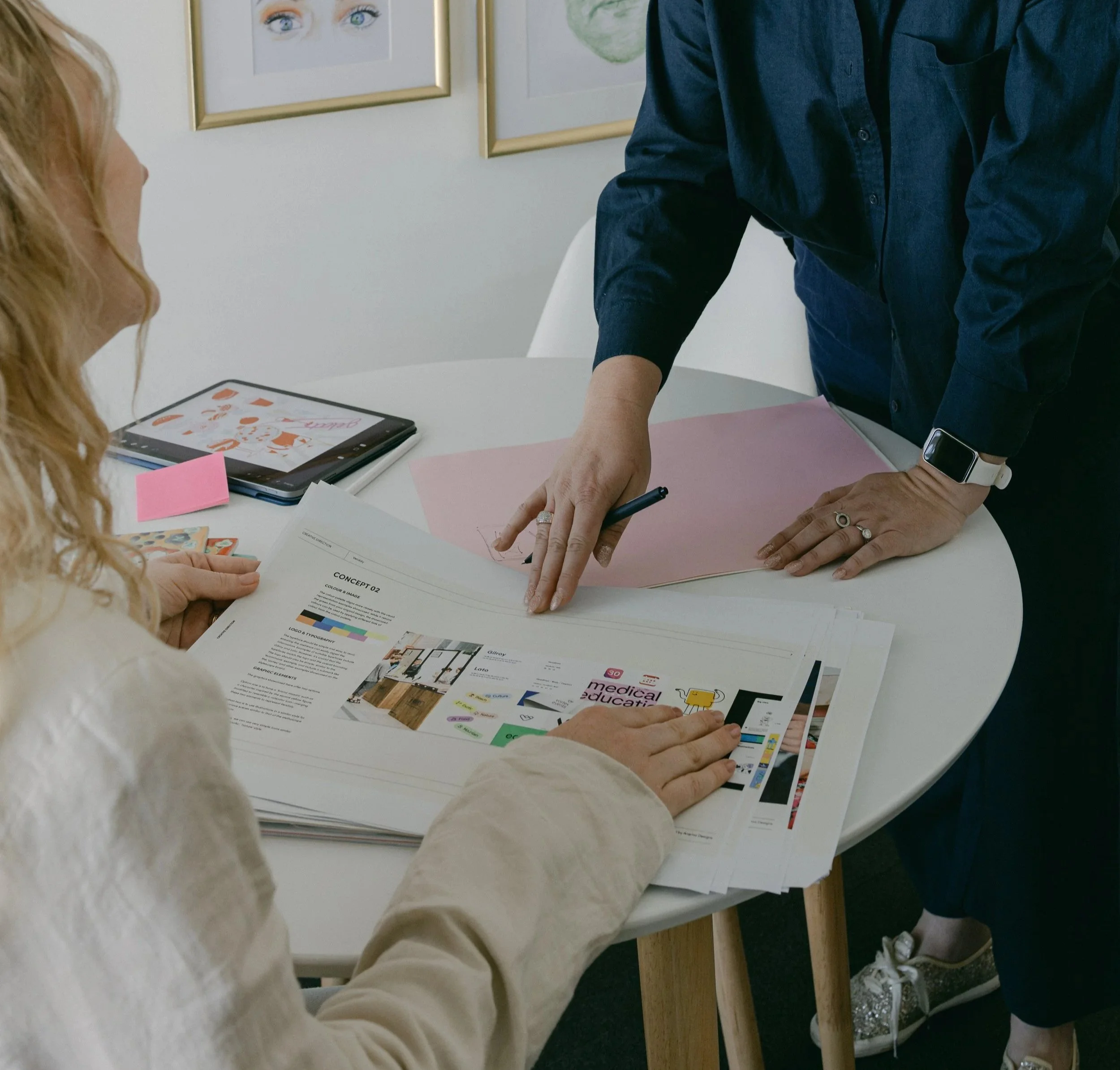Two women at a round table collaborating on design or project work, with documents, magazines, and a tablet visible.