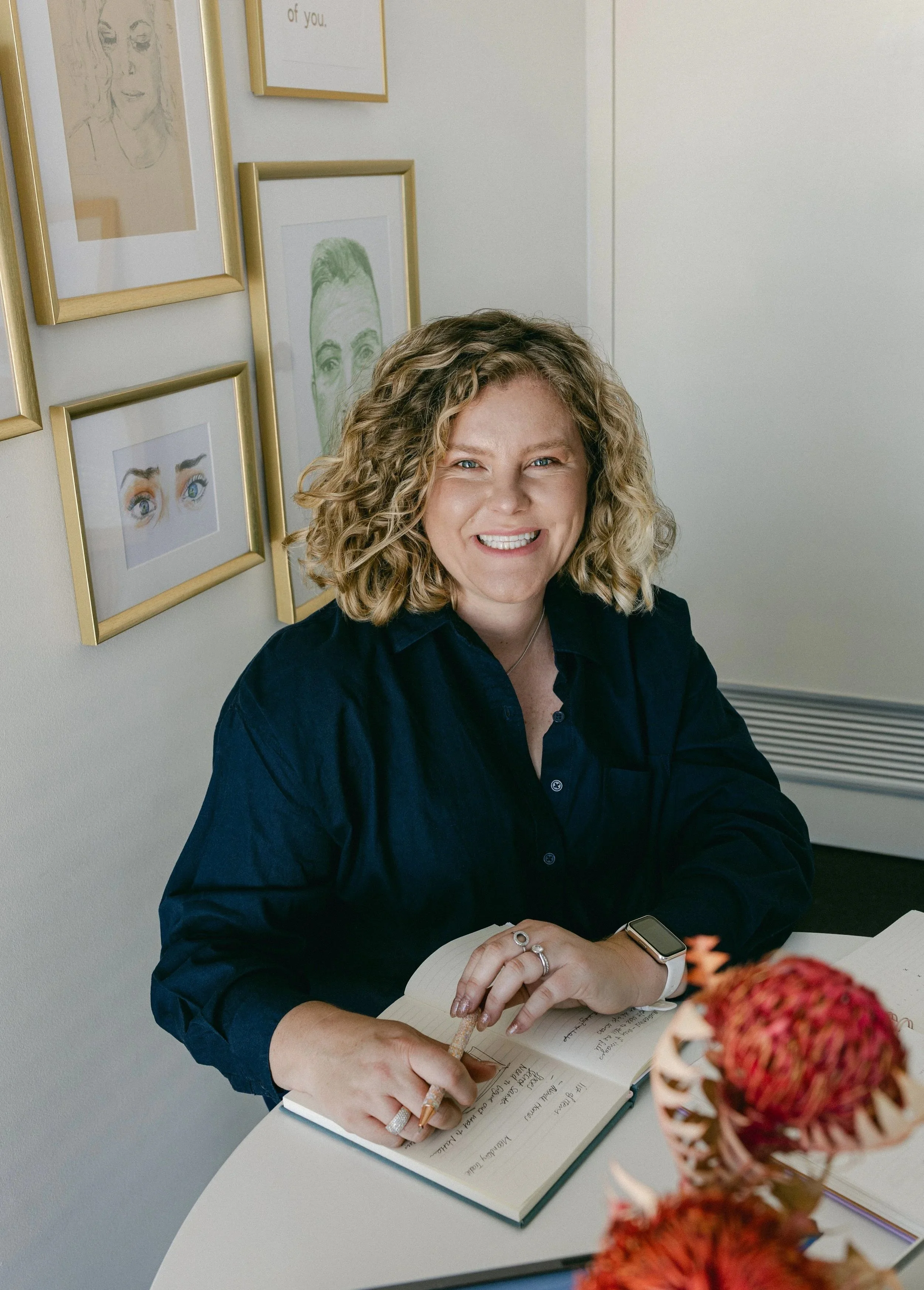 A woman with curly blonde hair smiling and writing in a notebook at a desk, with framed artwork on the wall behind her and a vase of orange flowers in the foreground.
