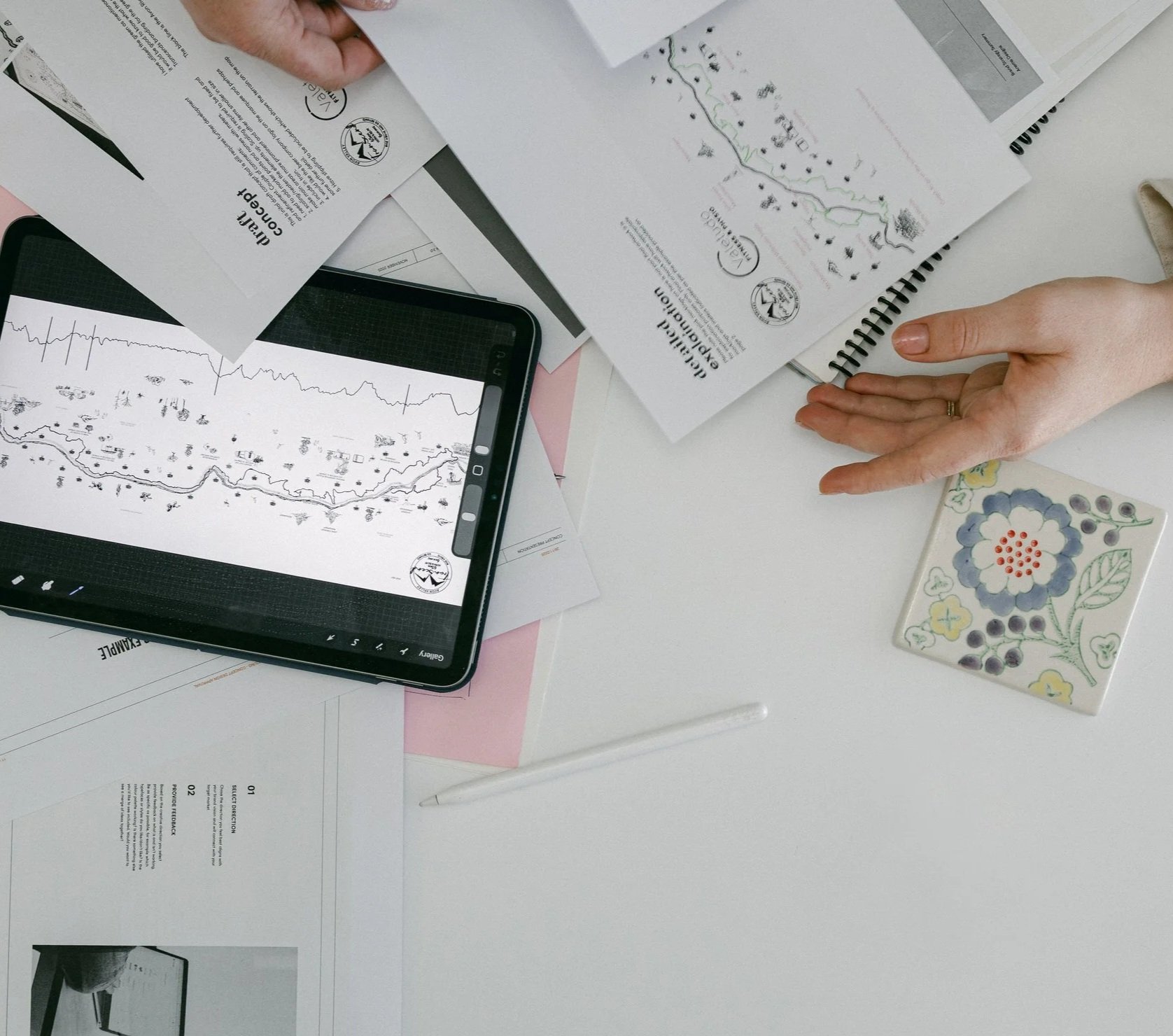 A top-down view of a workspace with scattered documents, a tablet displaying a map, a person's hand pointing, a decorative tile featuring a floral pattern, and a white pen on a white table.