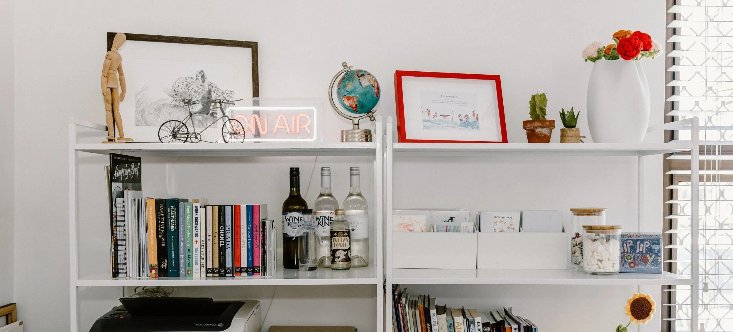 White bookshelf with books, glass bottles, framed art, plants, and decorative items in front of a window with blinds