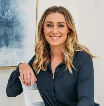 Portrait of a smiling woman with blonde hair, wearing a dark blue shirt, sitting in a well-lit room with artwork on the wall.