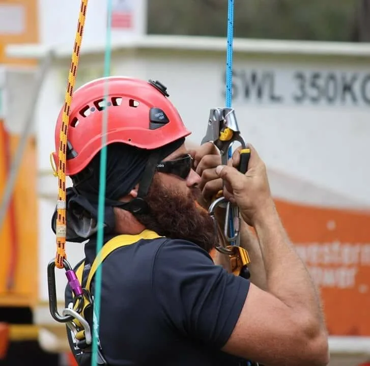 A man with a beard wearing a red helmet and black sunglasses adjusting his safety harness, with climbing gear hanging from his harness.