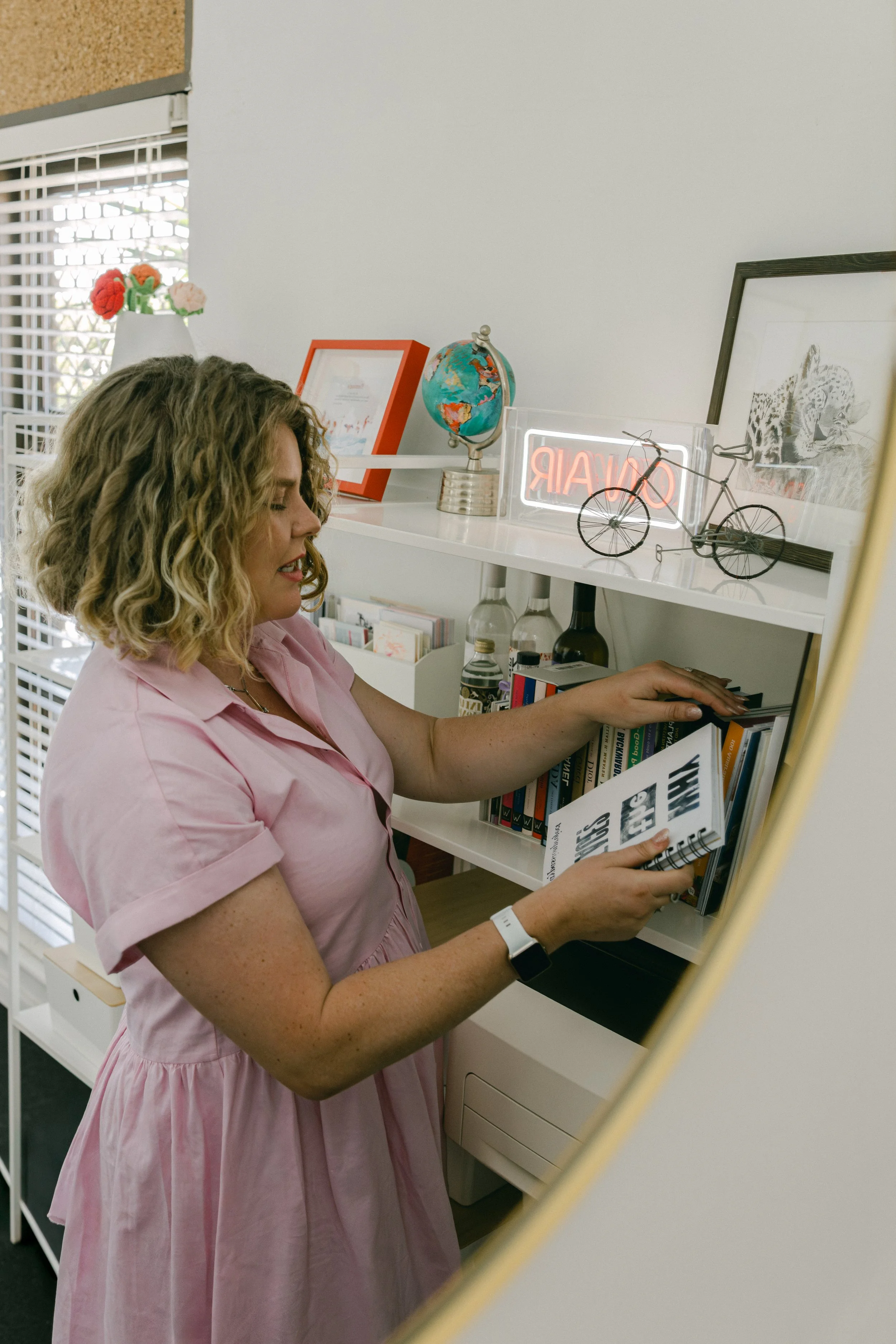 Woman with blonde curly hair wearing a pink dress, reading a book in a room with shelves containing decorative items like a globe and a bicycle sculpture.