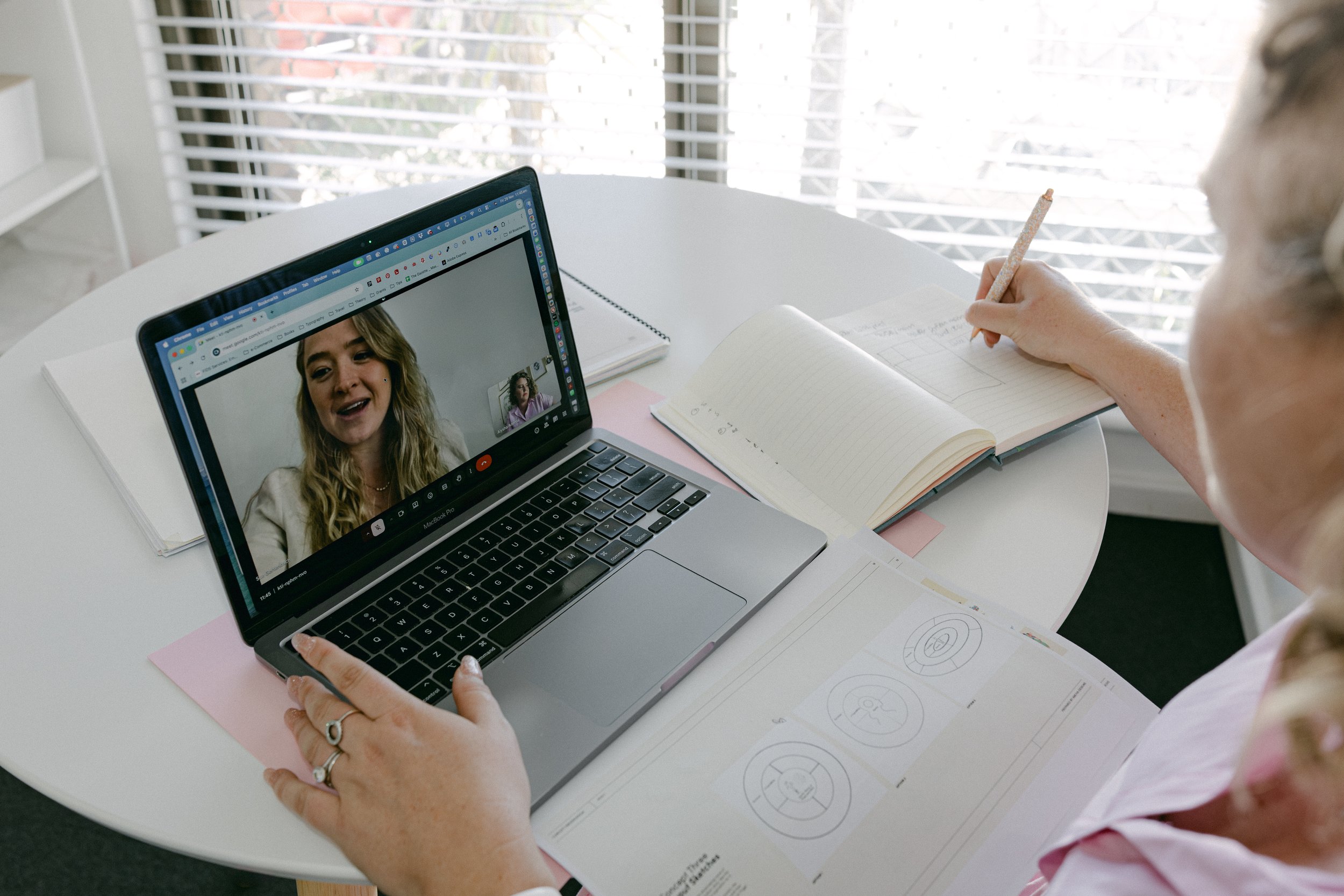 A woman sitting at a white table using a laptop for a video call with another woman. She has open notebooks and printed diagrams on the table, with blinds covering a window in the background.