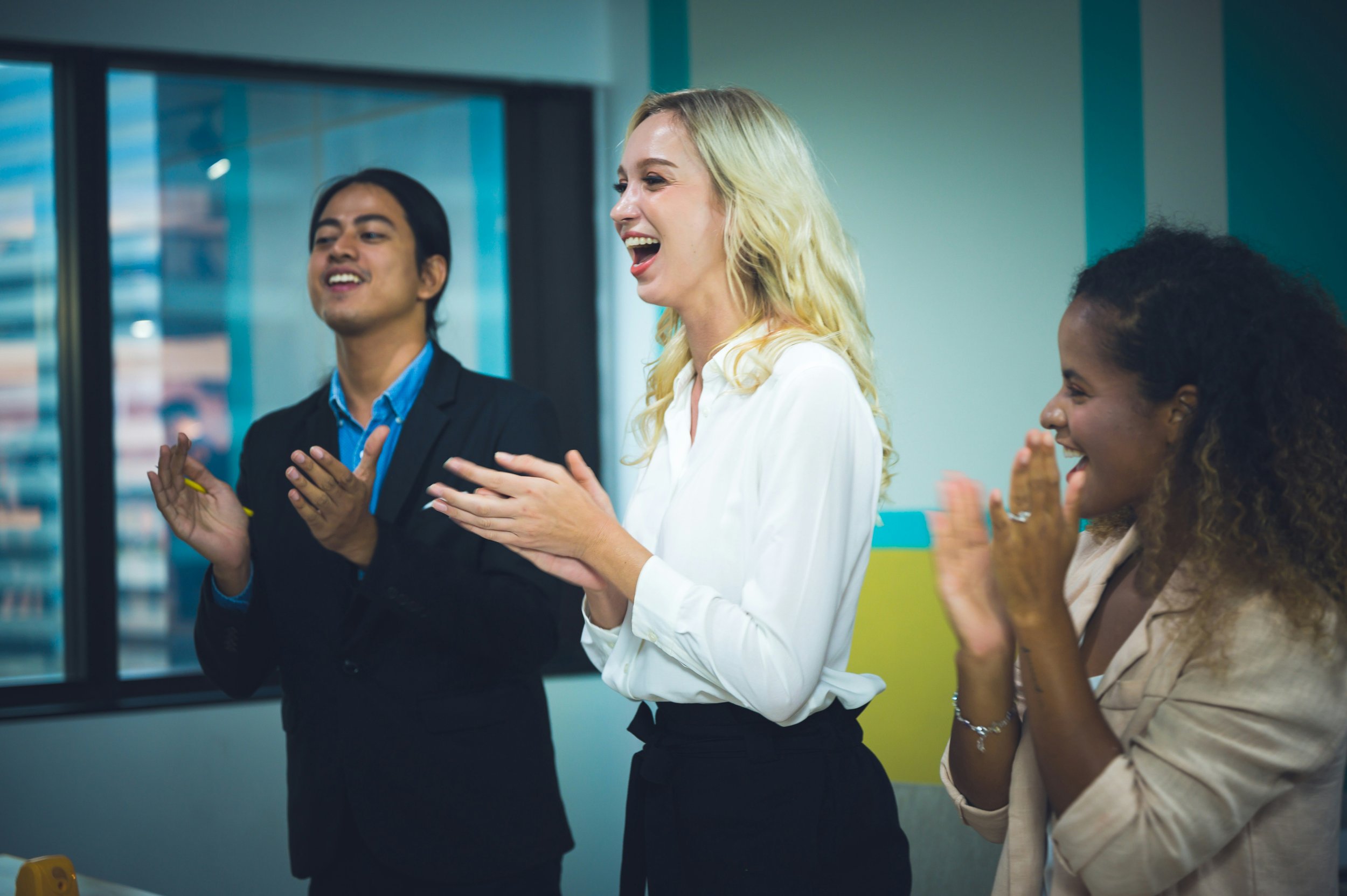 Three colleagues in a modern office smiling and clapping together, showing shared appreciation and team support.