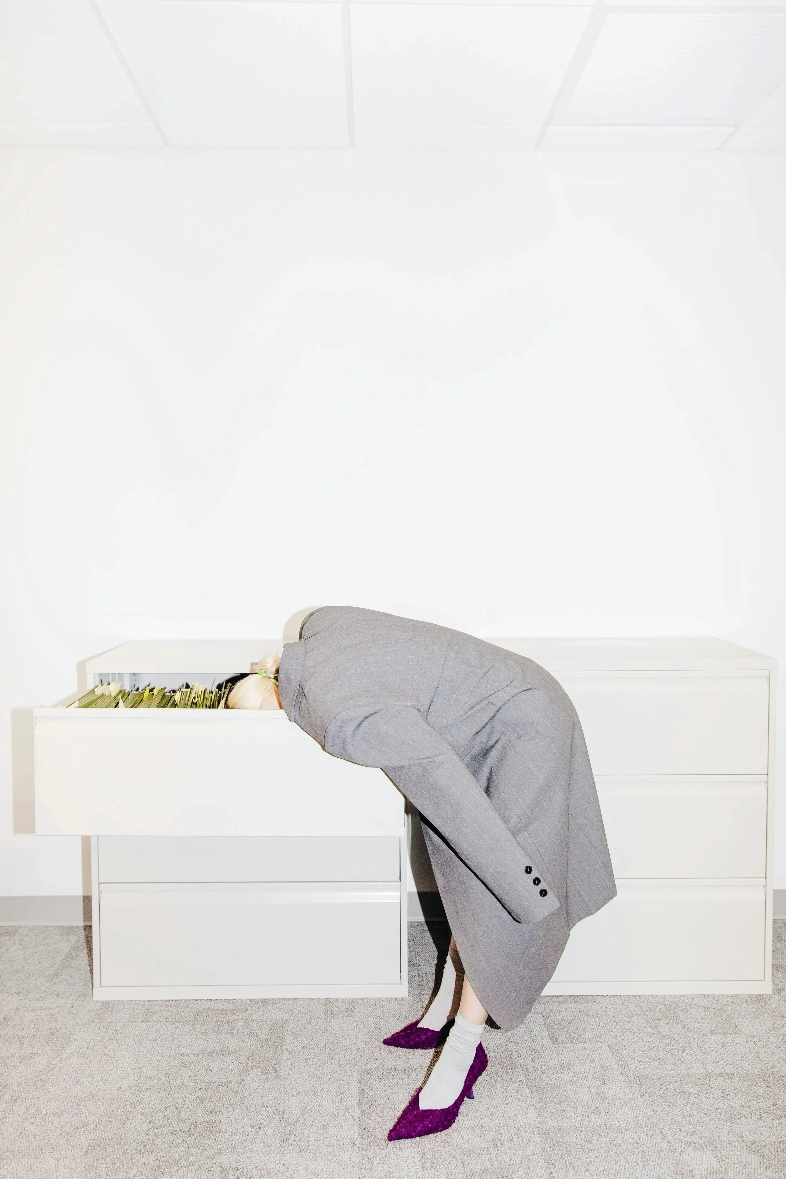 Artistic photograph of woman in grey coat and purple heels putting her head in a drawer of lettuce