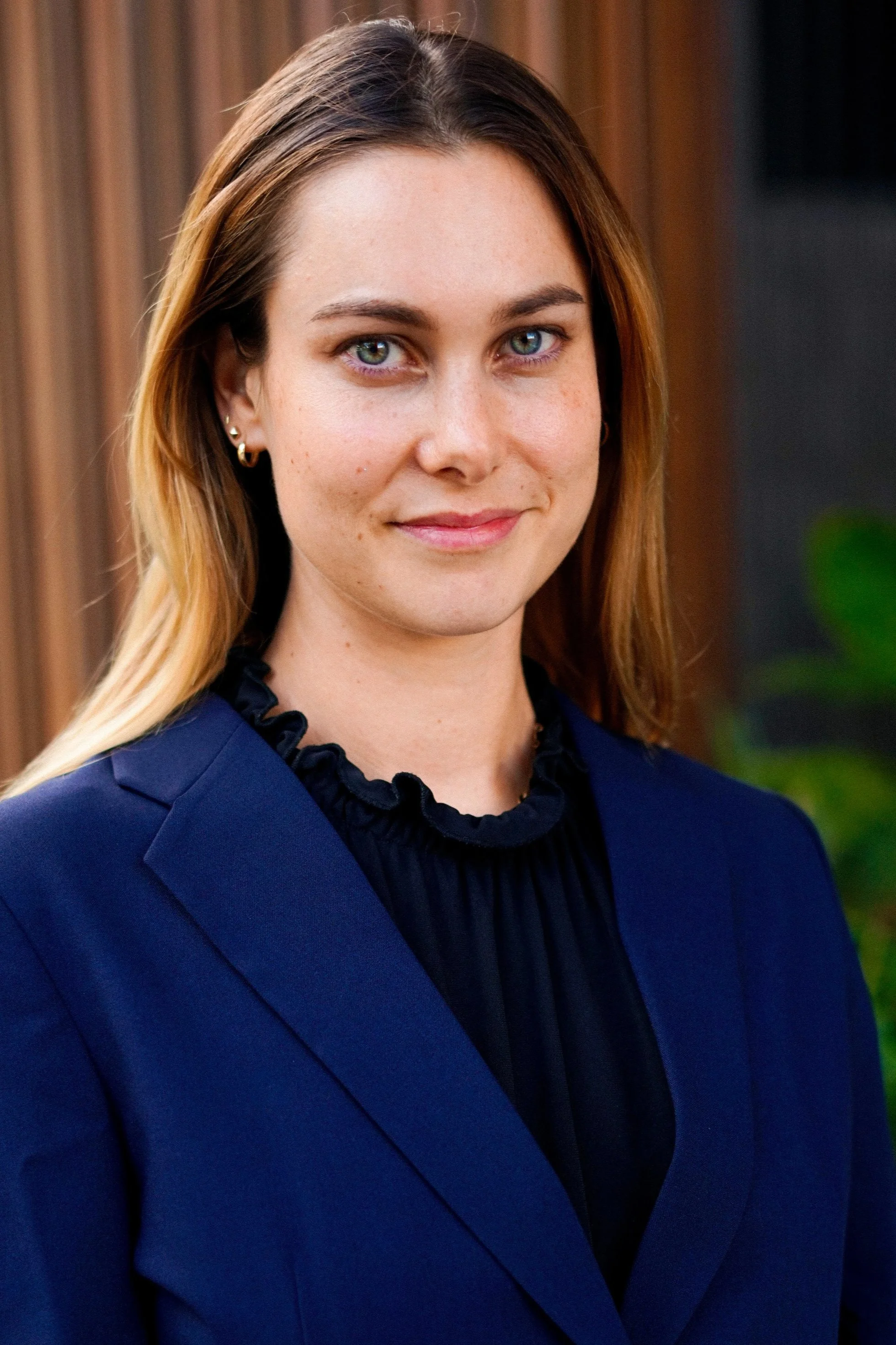 A woman with light skin, blue eyes, and long blonde hair is smiling softly at the camera. She is wearing a navy blue blazer and a black ruffled blouse, with small hoop earrings. The background features wooden paneling and a hint of green foliage.