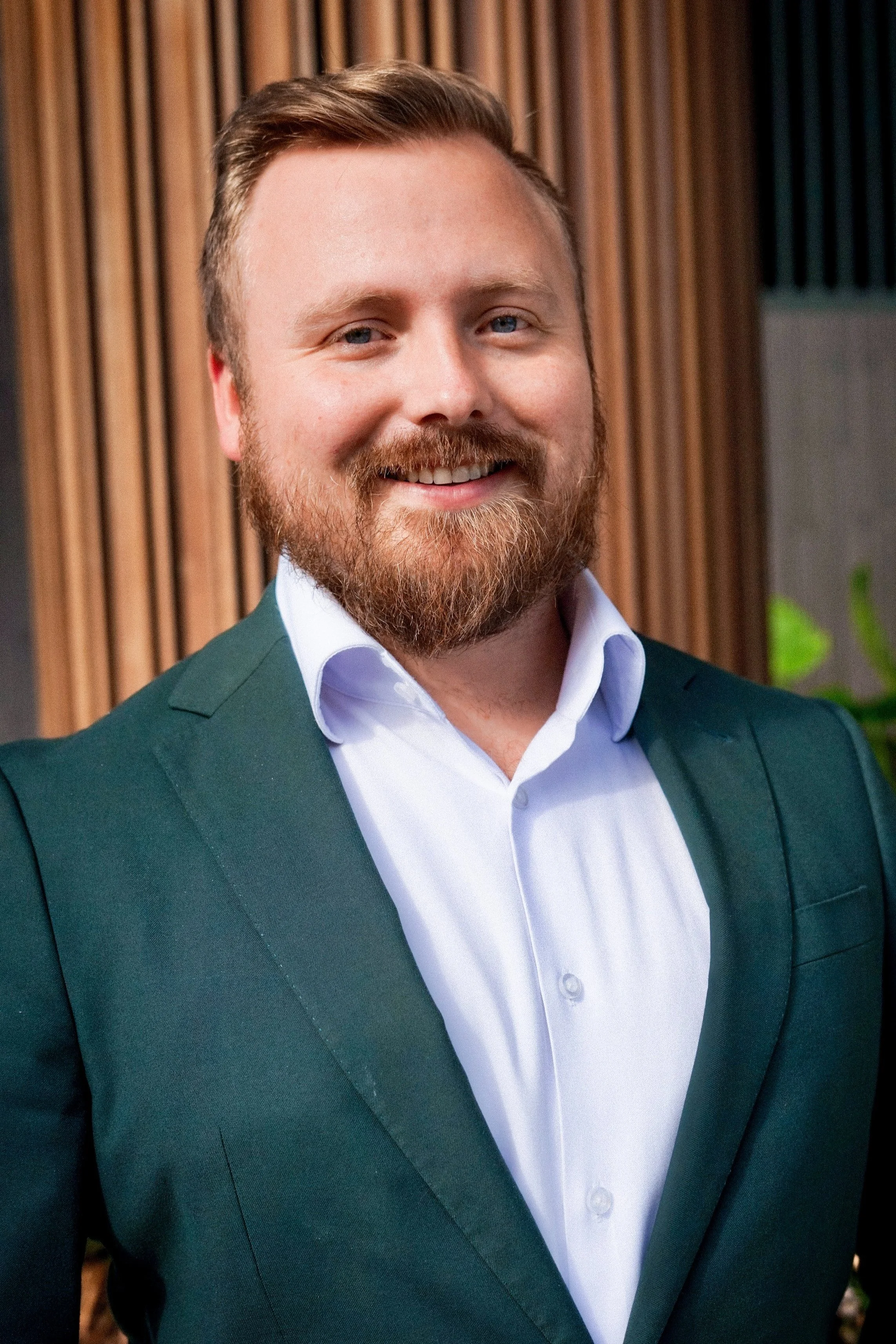 A smiling man with a beard and short hair, wearing a white dress shirt and a green blazer, standing in front of a wooden panel background.