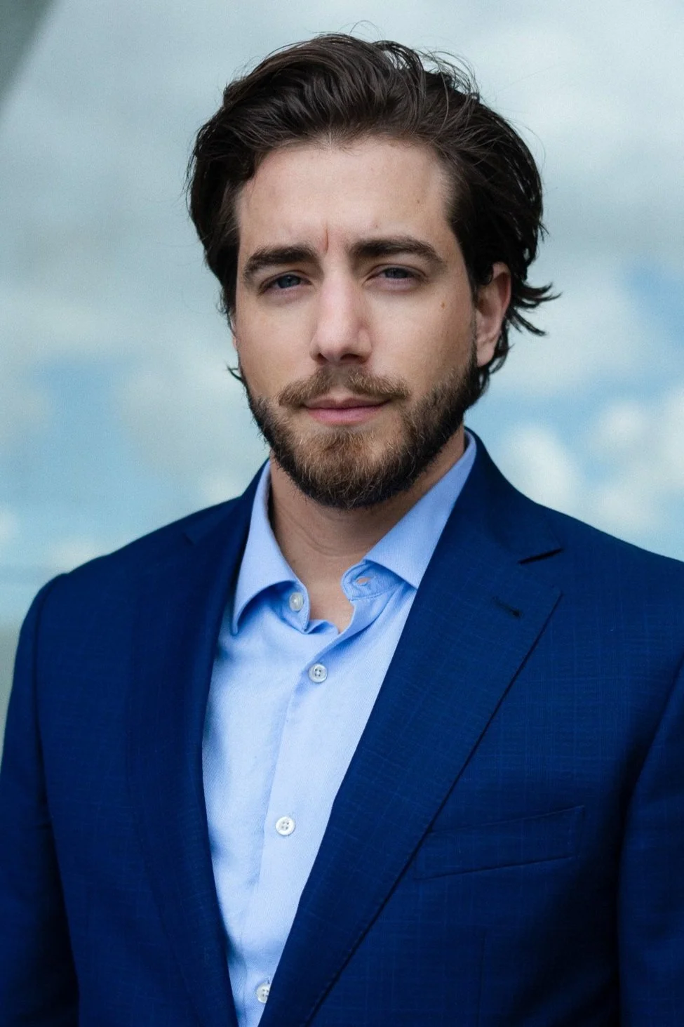 A young man with dark hair and a beard, dressed in a blue suit jacket and light blue shirt, stands outdoors with a cloudy sky in the background.