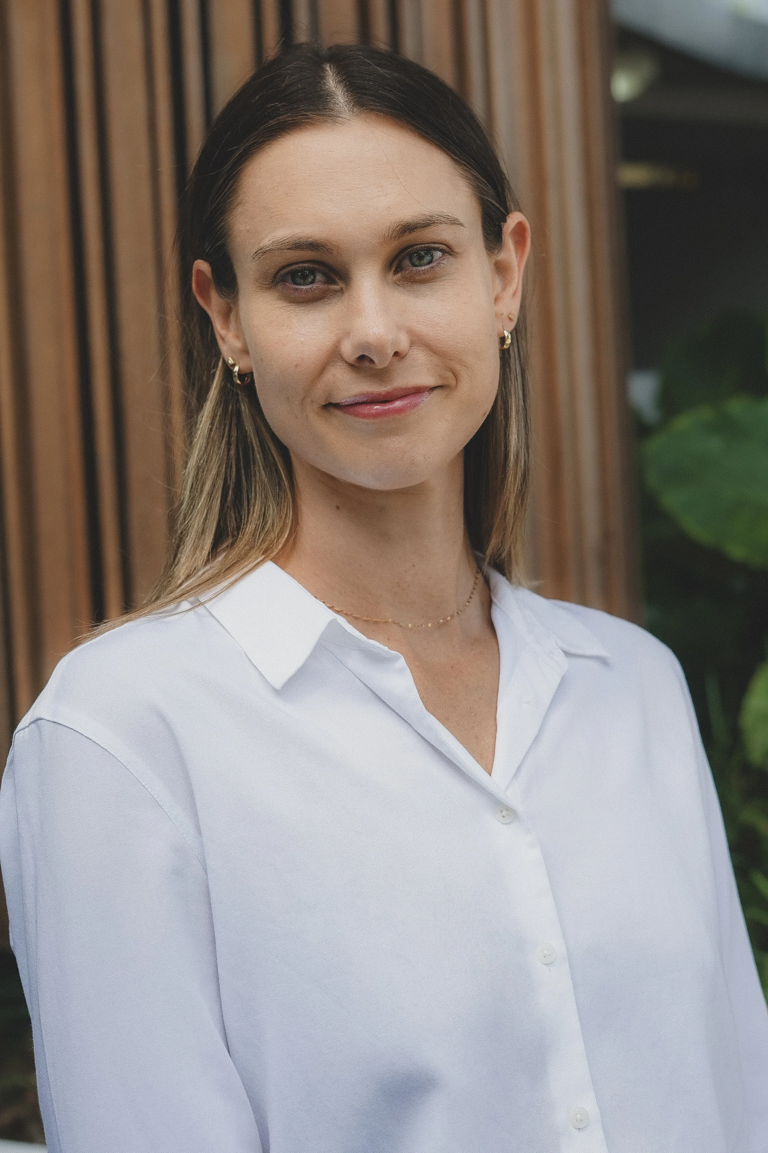 A woman with light skin, blue eyes, and long blonde hair is smiling softly at the camera. She is wearing a navy blue blazer and a black ruffled blouse, with small hoop earrings. The background features wooden paneling and a hint of green foliage.