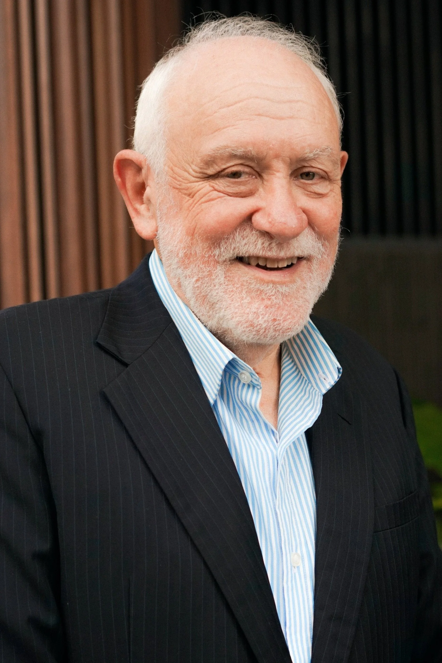 A smiling older man with a white beard and mustache, wearing a dark suit and a light blue collared shirt, standing in front of a background with wooden panels.