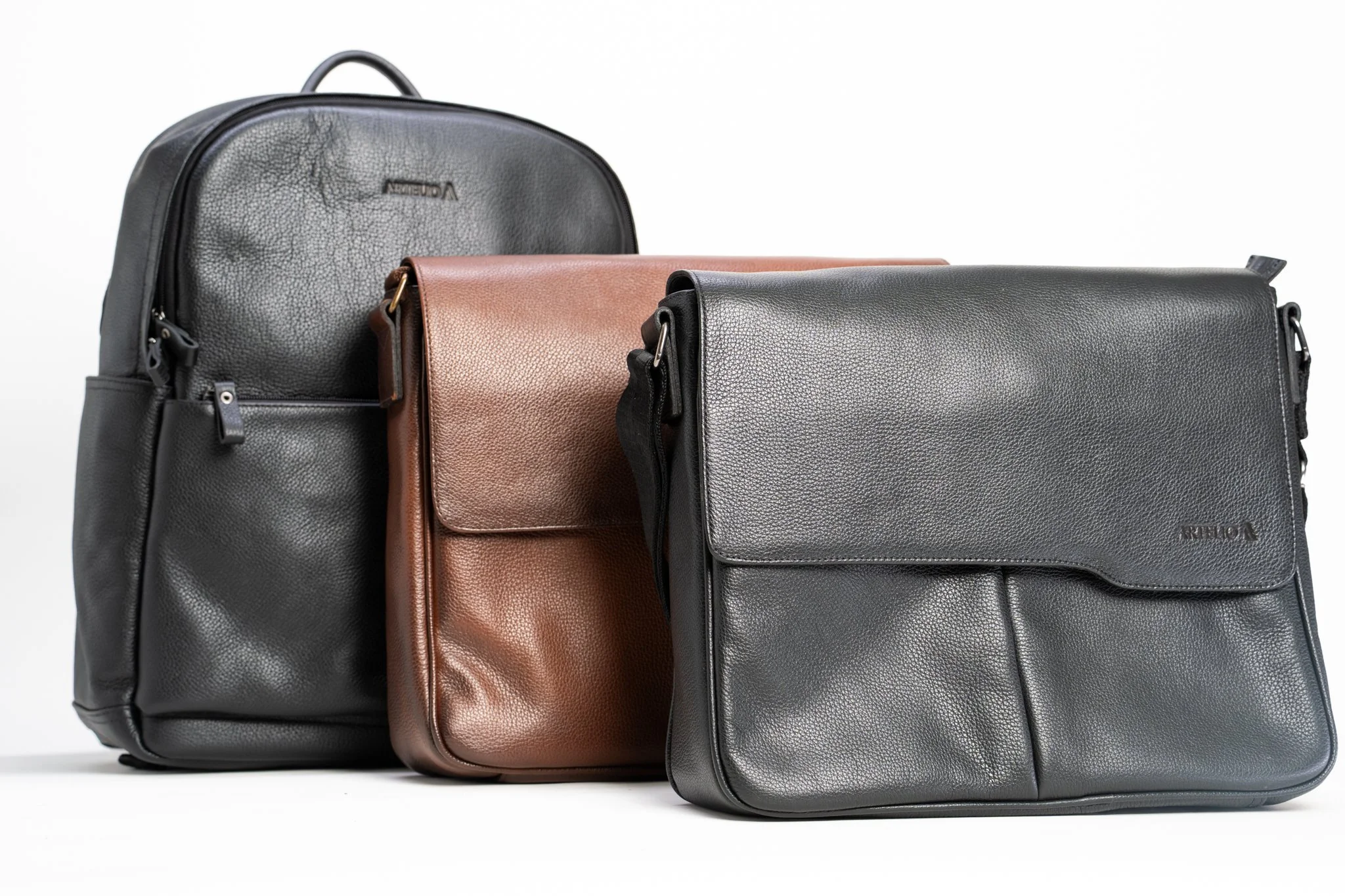 Three leather backpacks in black, brown, and gray, standing upright on a white background.
