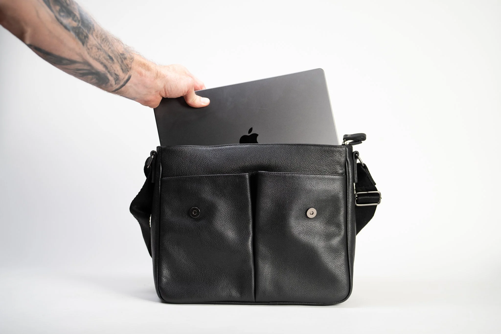 A person reaching into a black leather bag with multiple pockets, revealing a laptop with an Apple logo inside.