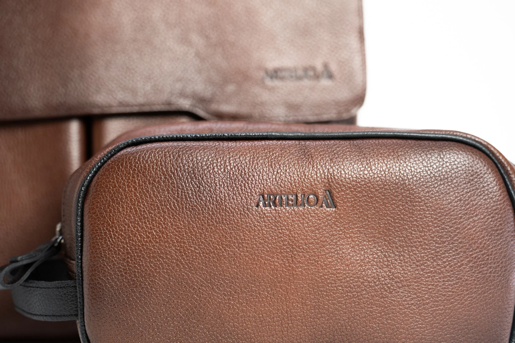 Close-up of a brown textured leather backpack with black piping, showing the brand name 'ARTELIOL' embossed on the front.
