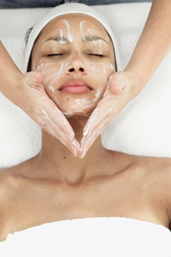 A woman receiving a facial massage while lying down with her eyes closed, her face covered in facial cream.