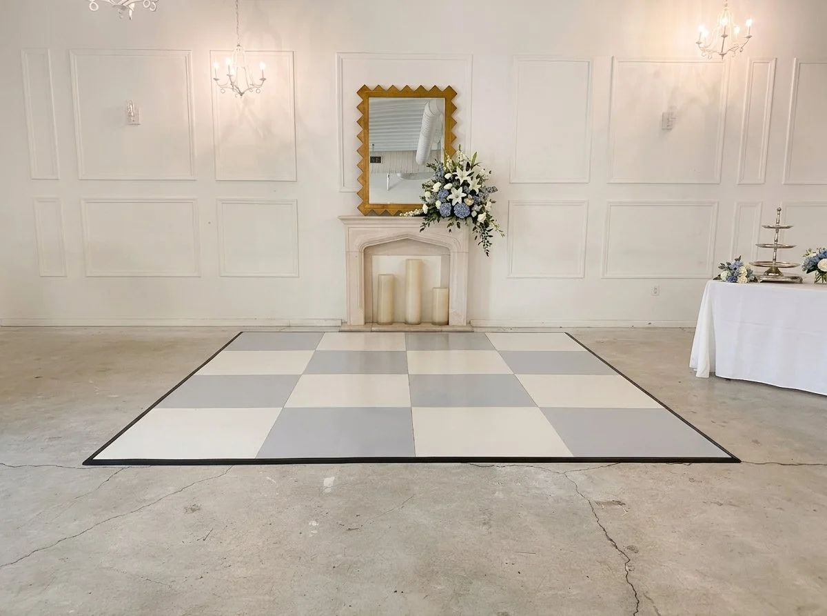 Detailed corner view of an ATL Dance Floors blue grey and white checkered dance floor panel in a refined parlor setting. Includes upholstered high-back chairs, an elegant stone fireplace with candles, and a vintage gold-framed mirror, demonstrating u
