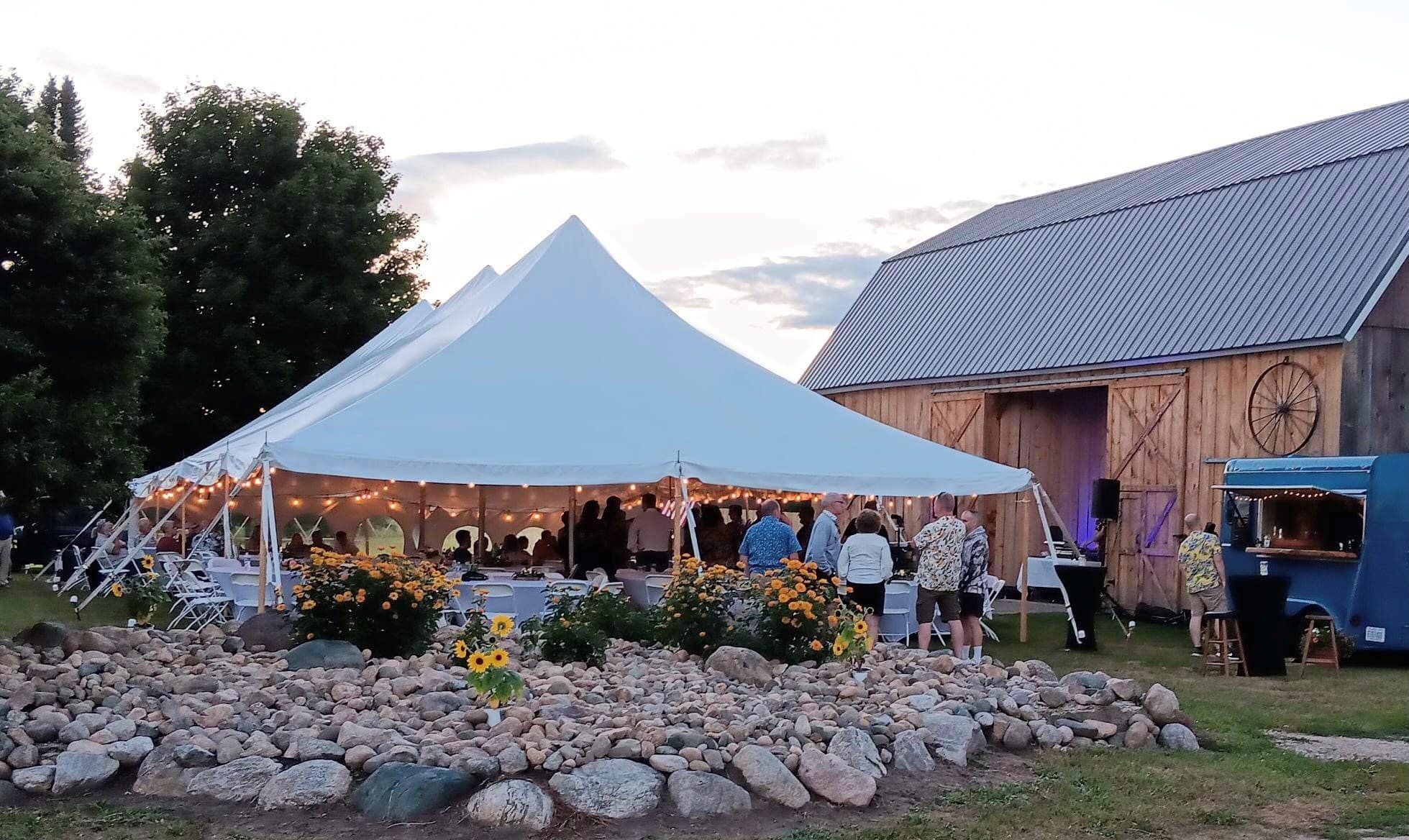 Classic pole tent set up for an outdoor wedding reception near a barn, with open sides, café lighting, and guest seating in a natural outdoor setting.