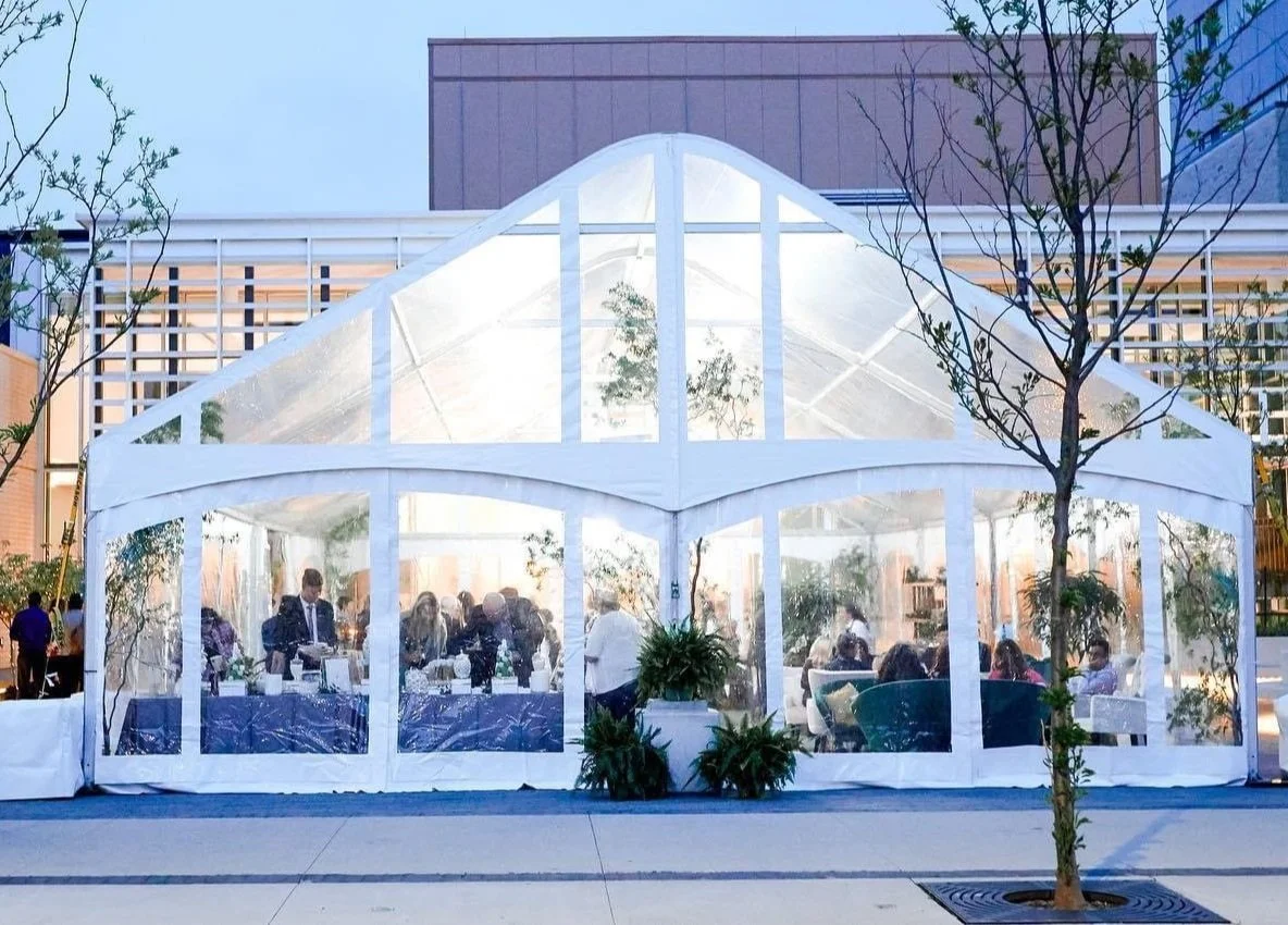 A large white tent set up outdoors with transparent sides, filled with people sitting and chatting, surrounded by small decorative trees and plants, with multi-story buildings in the background.