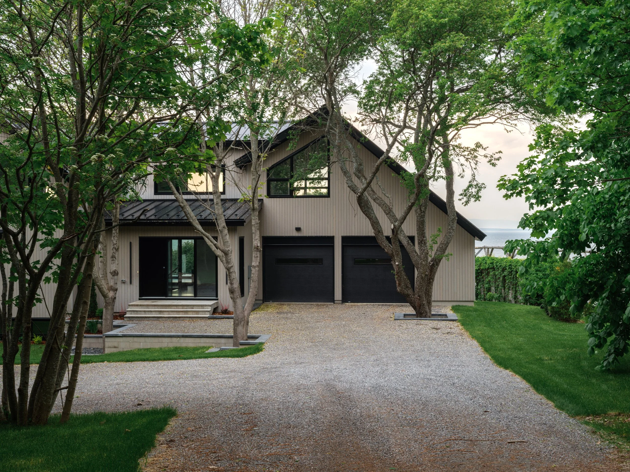Maison moderne en bois beige avec deux garages noirs, entourée d'arbres et de verdure, avec vue sur un lac en arrière-plan.