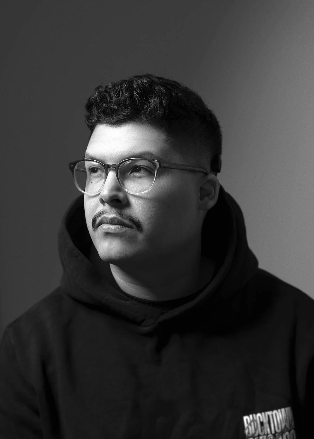 Black and white portrait of a man with glasses, a mustache, wearing a hoodie, looking to the side.