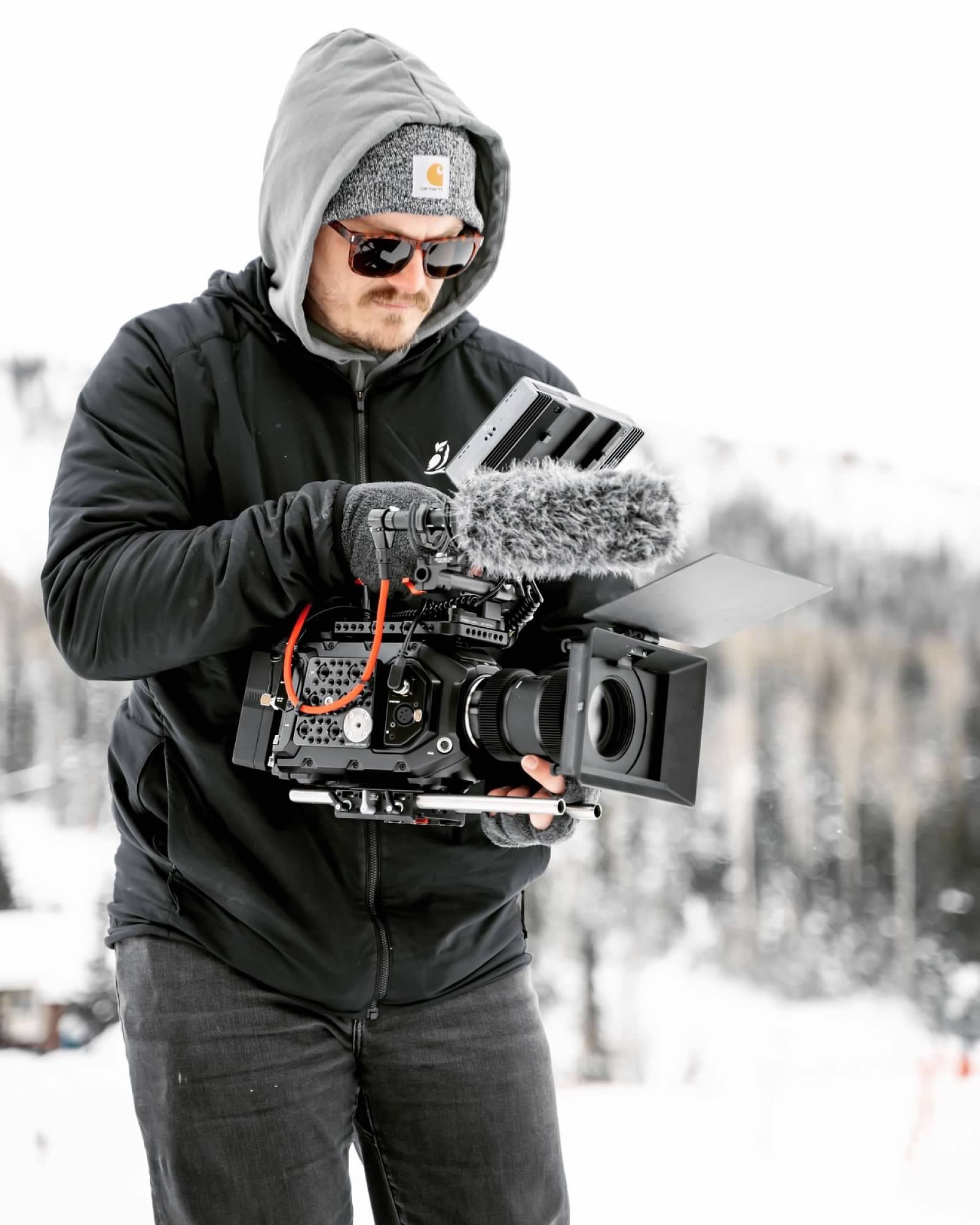 Man dressed in winter clothing operating a professional film camera with a furry microphone attached outdoors in snowy weather.