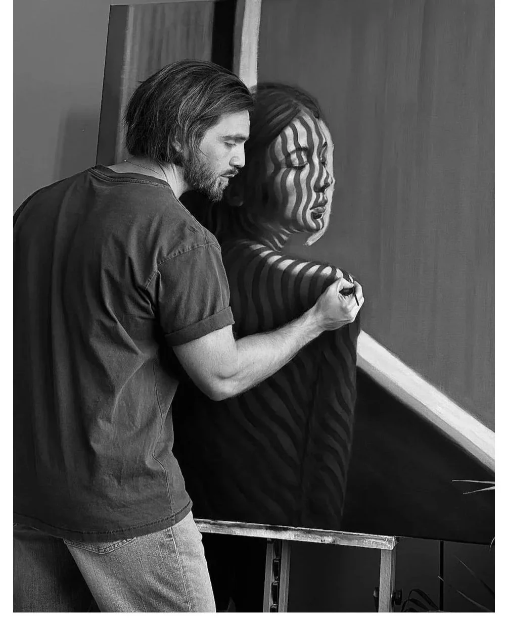 A male artist painting a woman's face on a large canvas, using black and white tones, in an art studio.