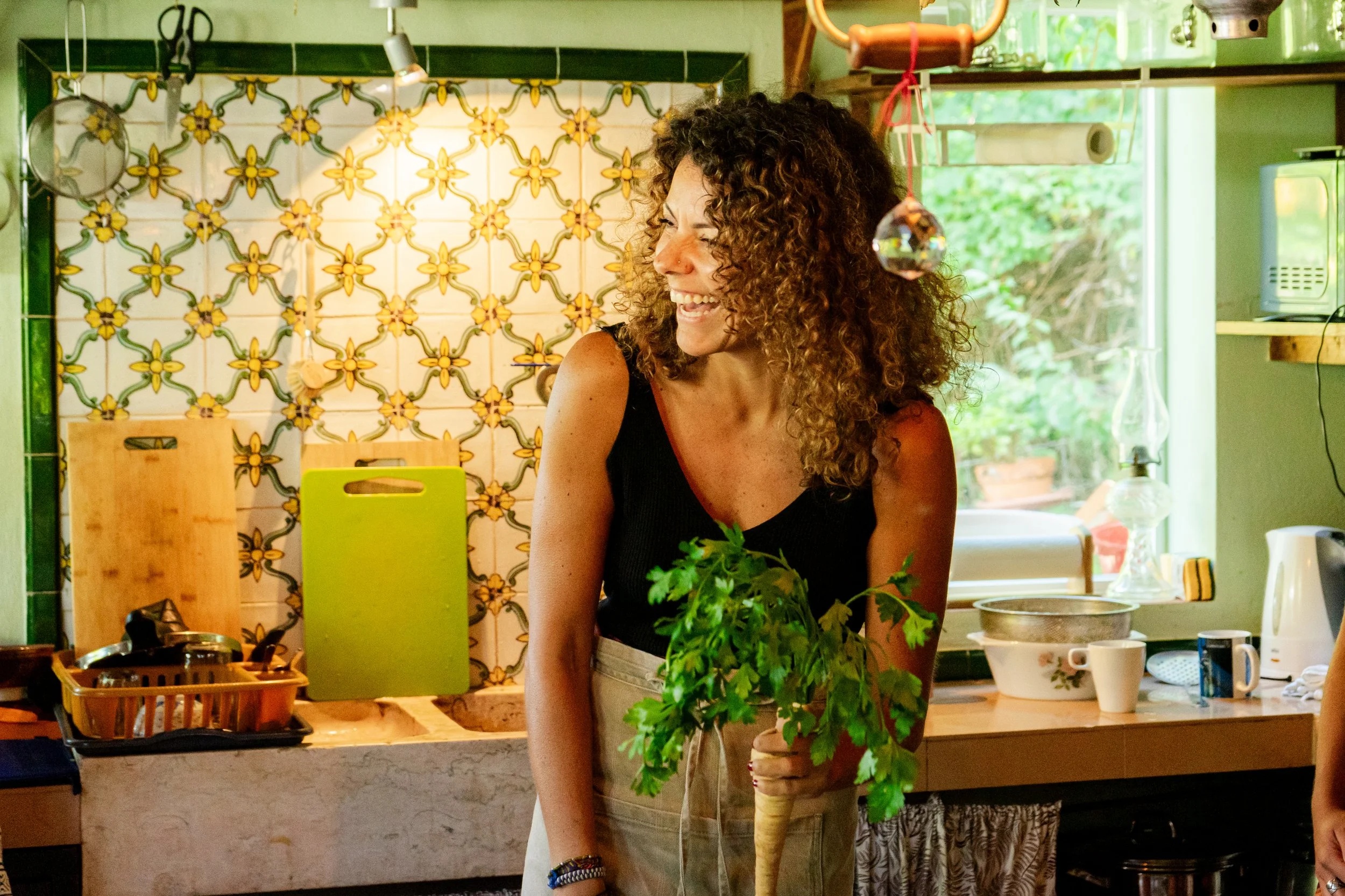 Mulher sorridente segurando uma cenoura com salsa na cozinha, decorada com azulejos coloridos e utensílios de cozinha ao fundo.