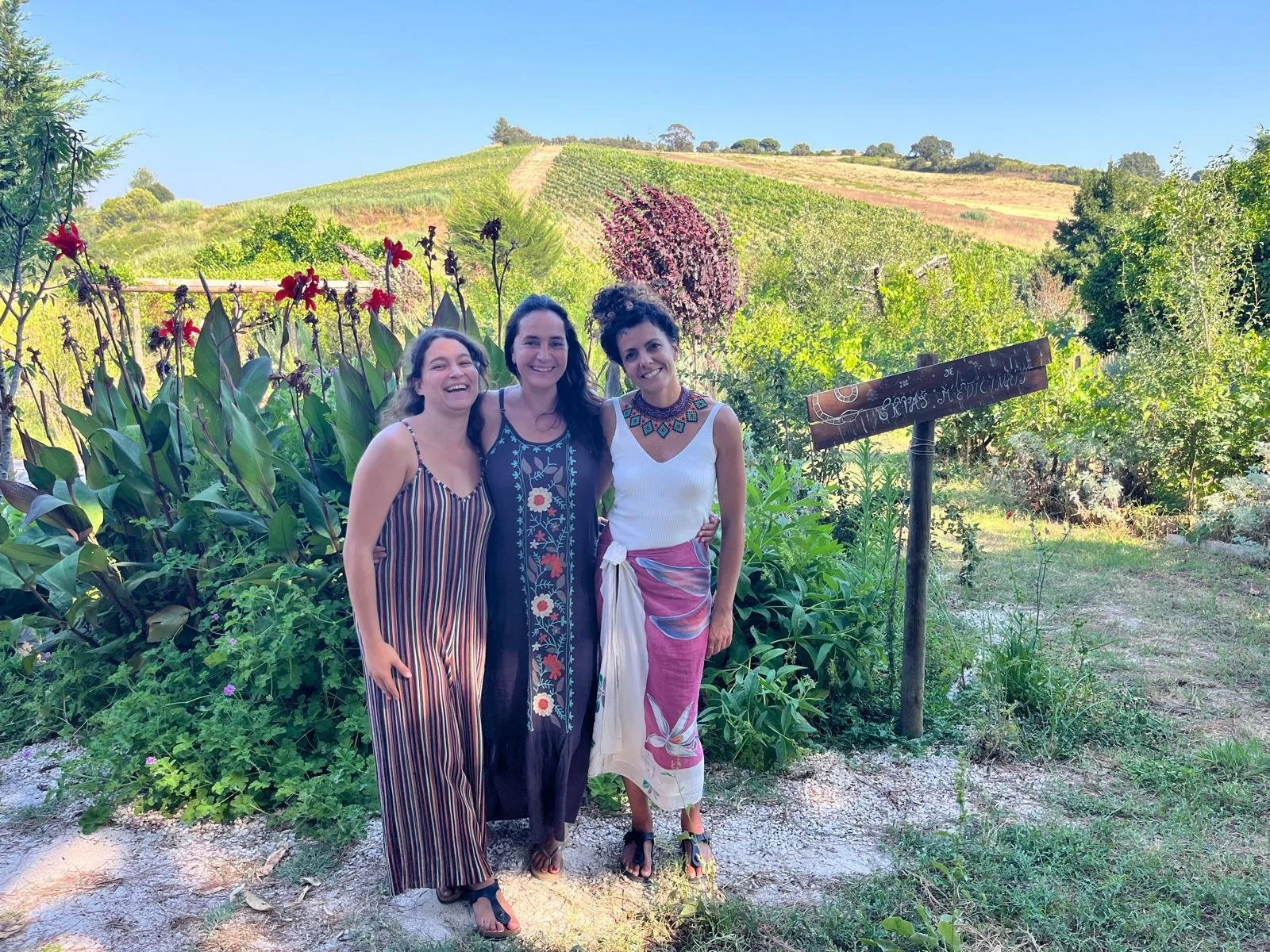 Três mulheres sorrindo em um jardim com plantas e flores, com colinas verdes ao fundo e céu claro.