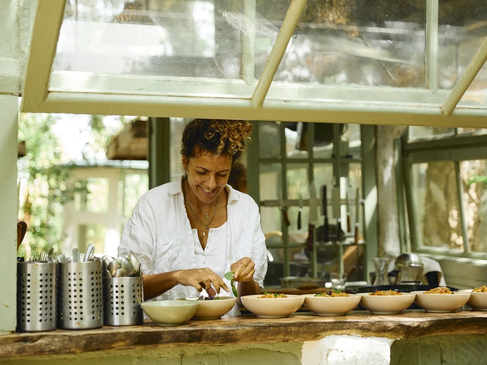 Mulher sorridente ao preparar alimentos em uma cozinha com janelas grandes e muita luz natural.