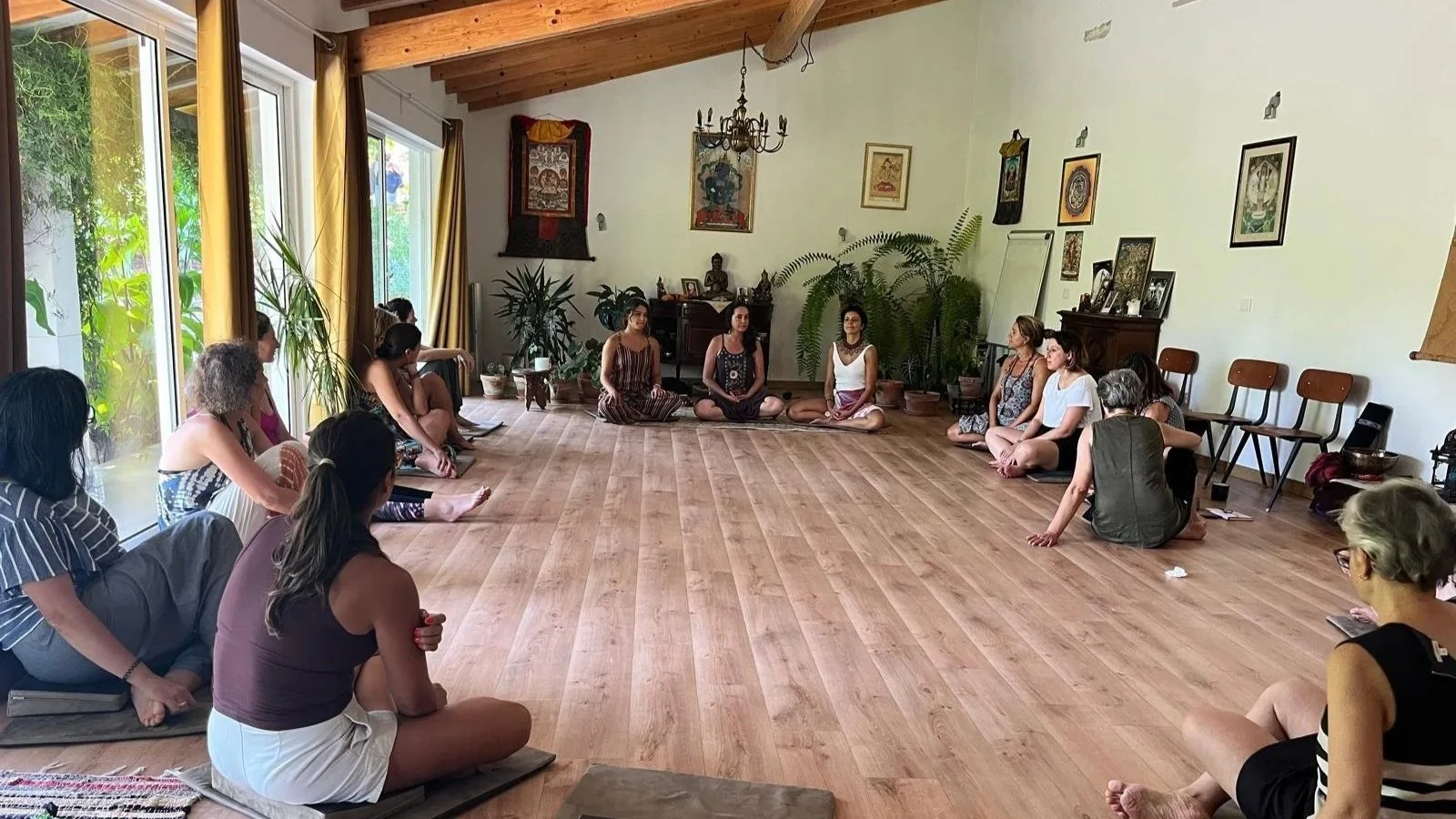 Grupo de pessoas sentadas em círculo no chão de um salão, participando de uma atividade de meditação ou yoga, com plantas e quadros na parede ao fundo.