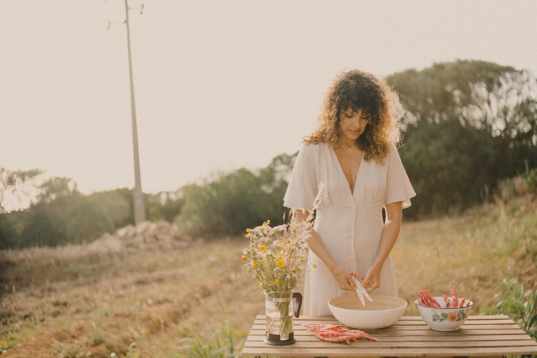 Mulher com cabelo cacheado morena vestindo vestido claro cortando carne em uma mesa ao ar livre, com um buquê de flores ao lado e uma tigela com legumes.