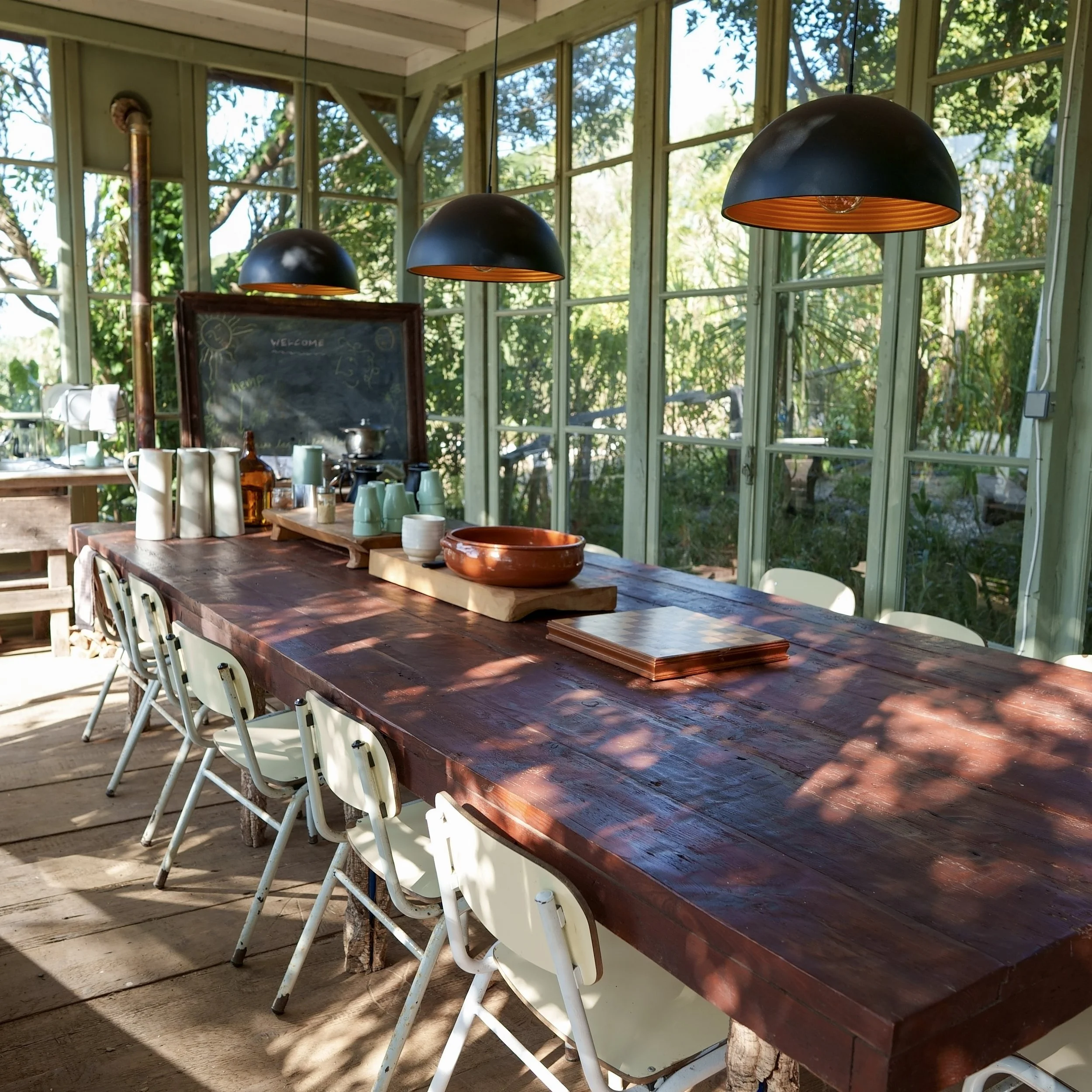Sala de jantar com mesa de madeira rústica, várias cadeiras brancas, janelas de vidro, iluminação suspensa com lâmpadas pretas, objetos de cozinha e quadro negro com escrita