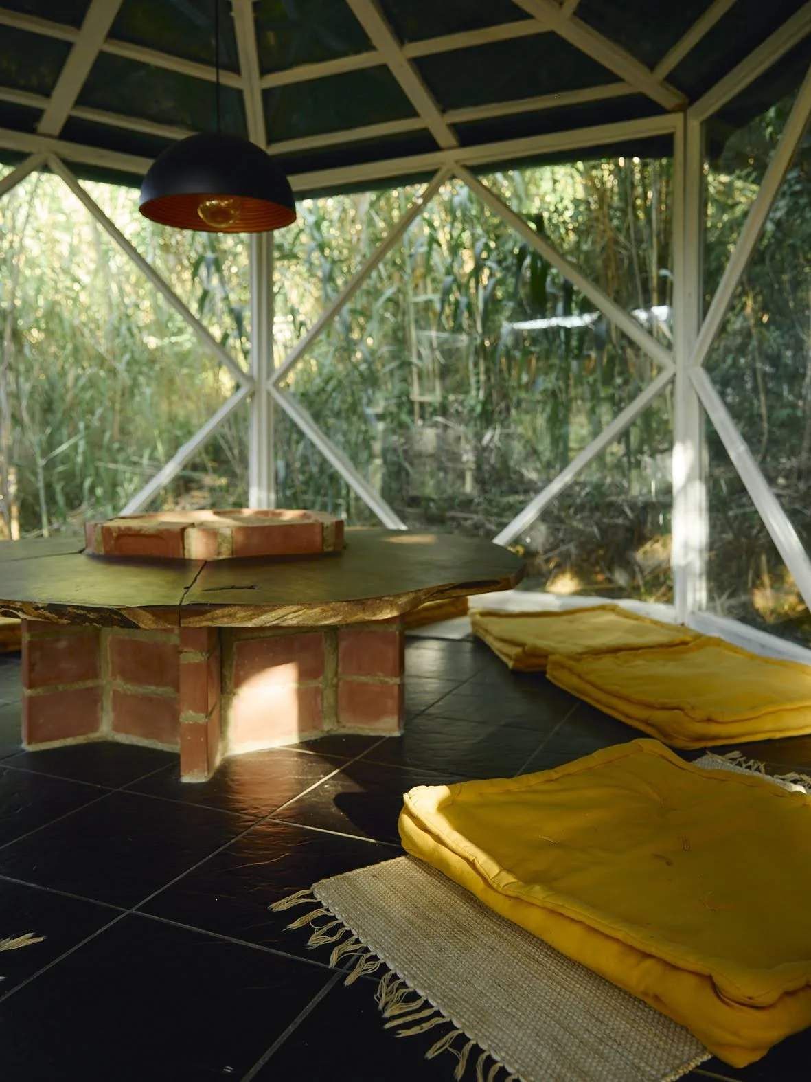 Interior de uma cabana de vidro com uma mesa de centro de madeira, almofadas amarelas no chão e um lustre preto.