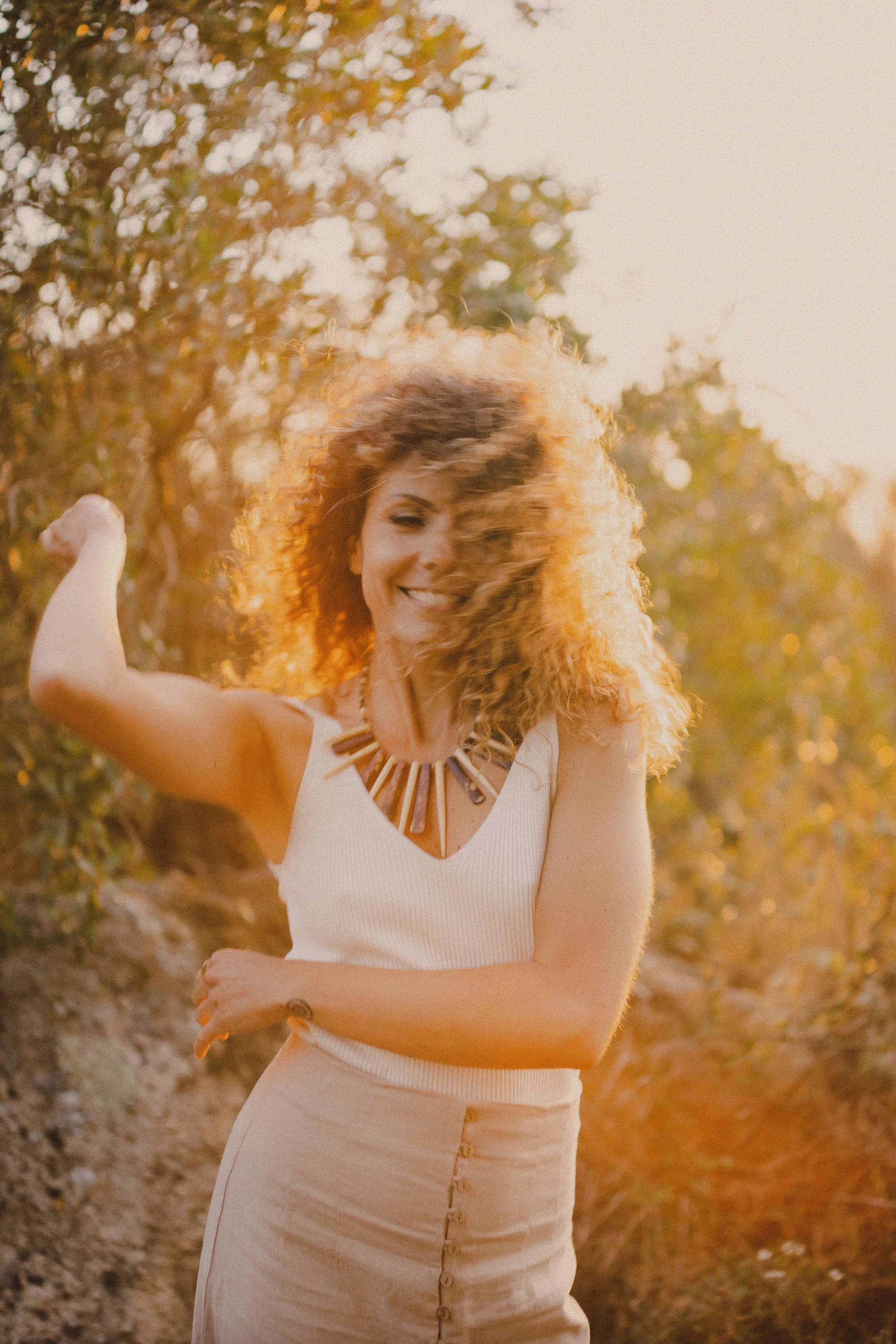 Mulher sorridente com cabelos cacheados ao entardecer em ambiente ao ar livre, vestindo blusa branca e saia bege, usando colar de madeira, com fundo de árvores.