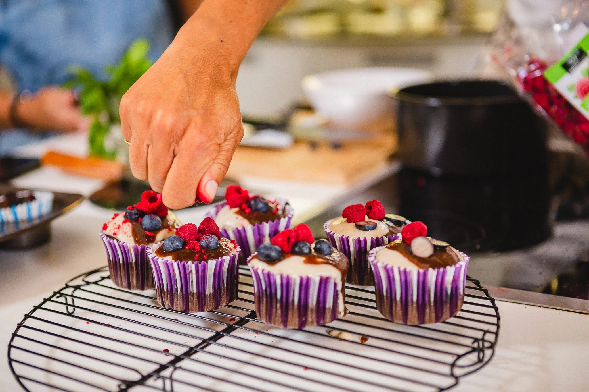 Cupcakes decorados com frutas vermelhas e blueberries, dispostos sobre uma grade de confeitaria em uma cozinha.